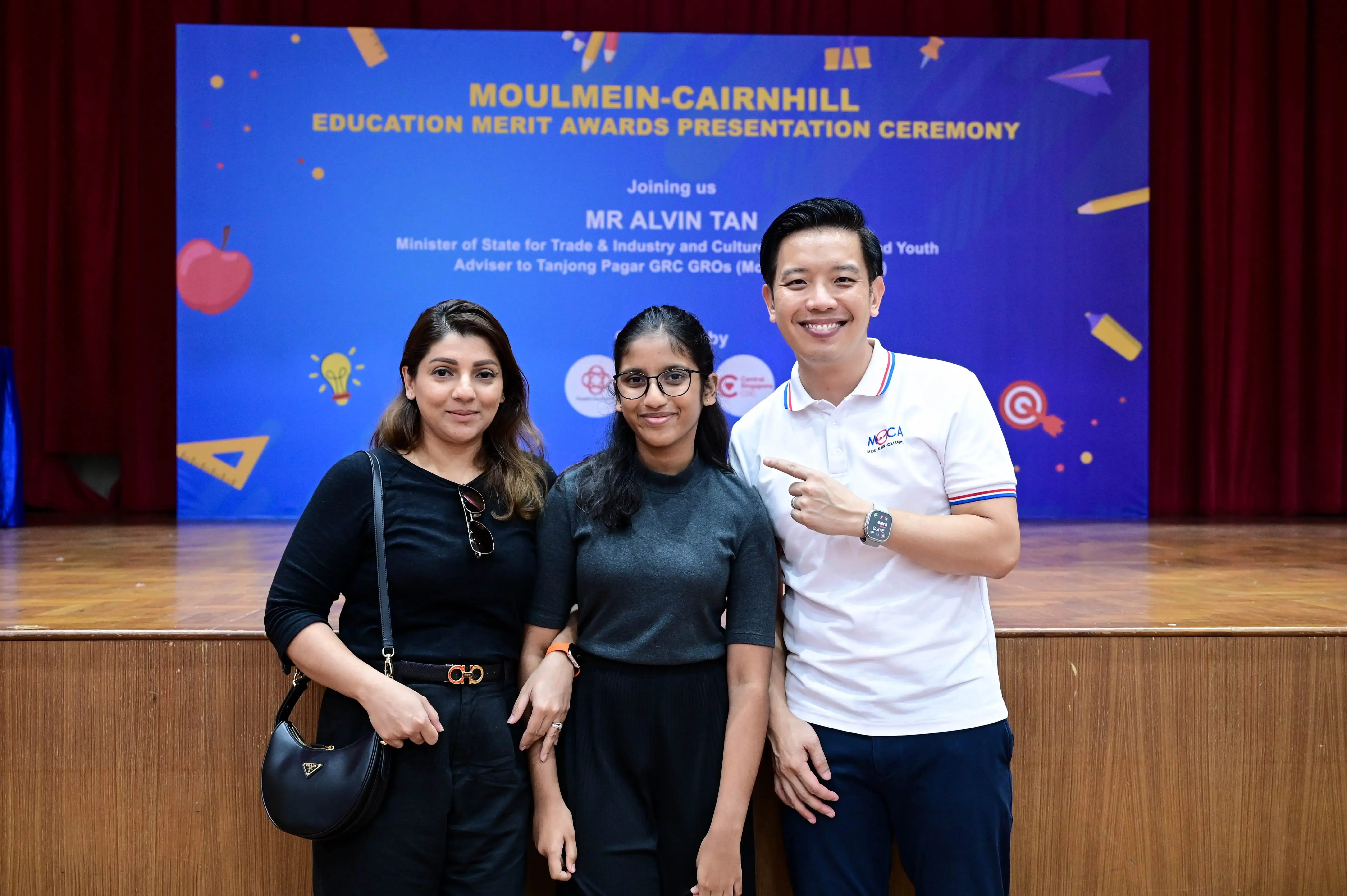 Three people smile in front of a blue Moulmein-Cairnhill Education Merit Awards Presentation Ceremony banner.
