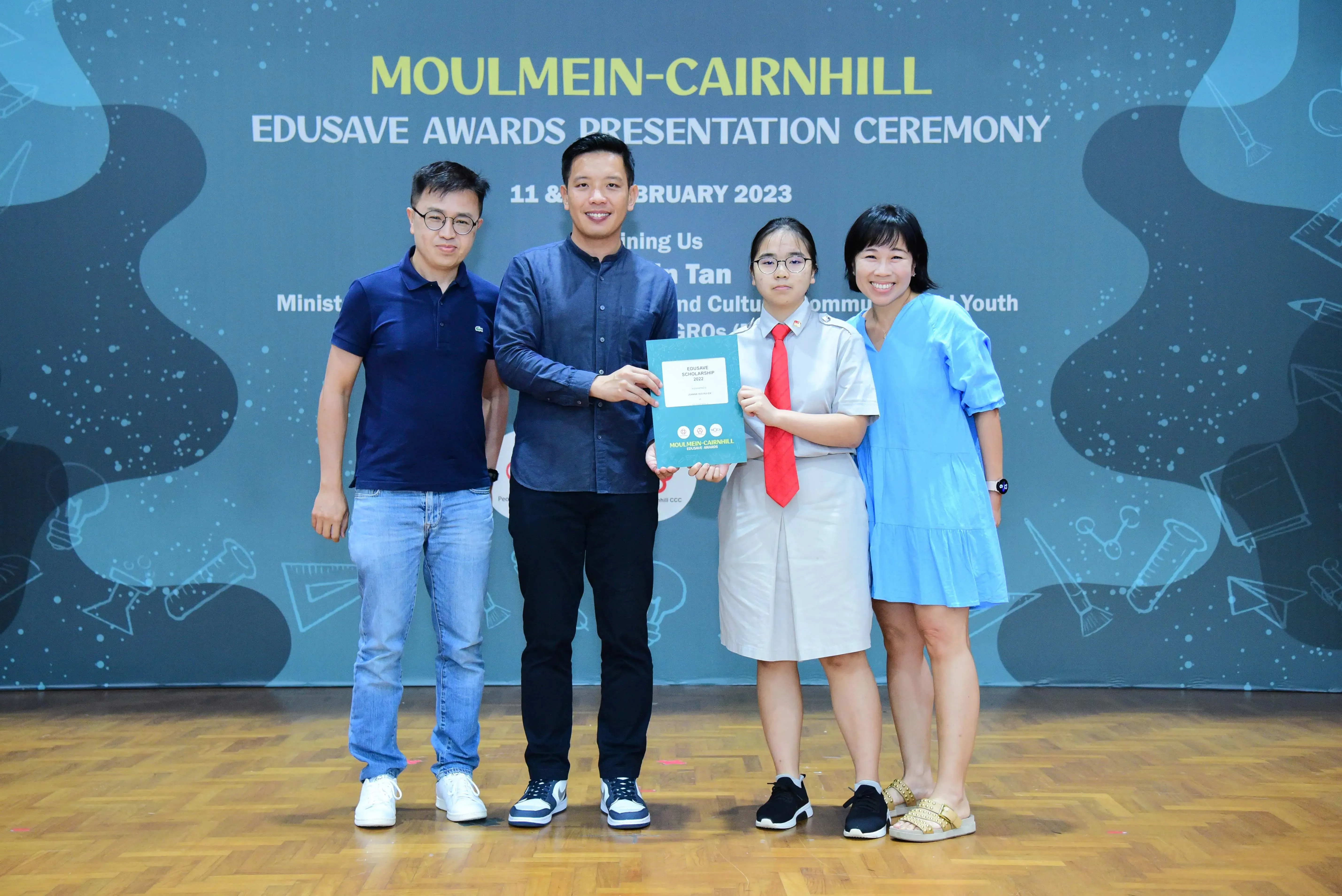 Four people pose on stage at the Moulmein-Cairnhill Edusave Awards Presentation Ceremony, holding a certificate.