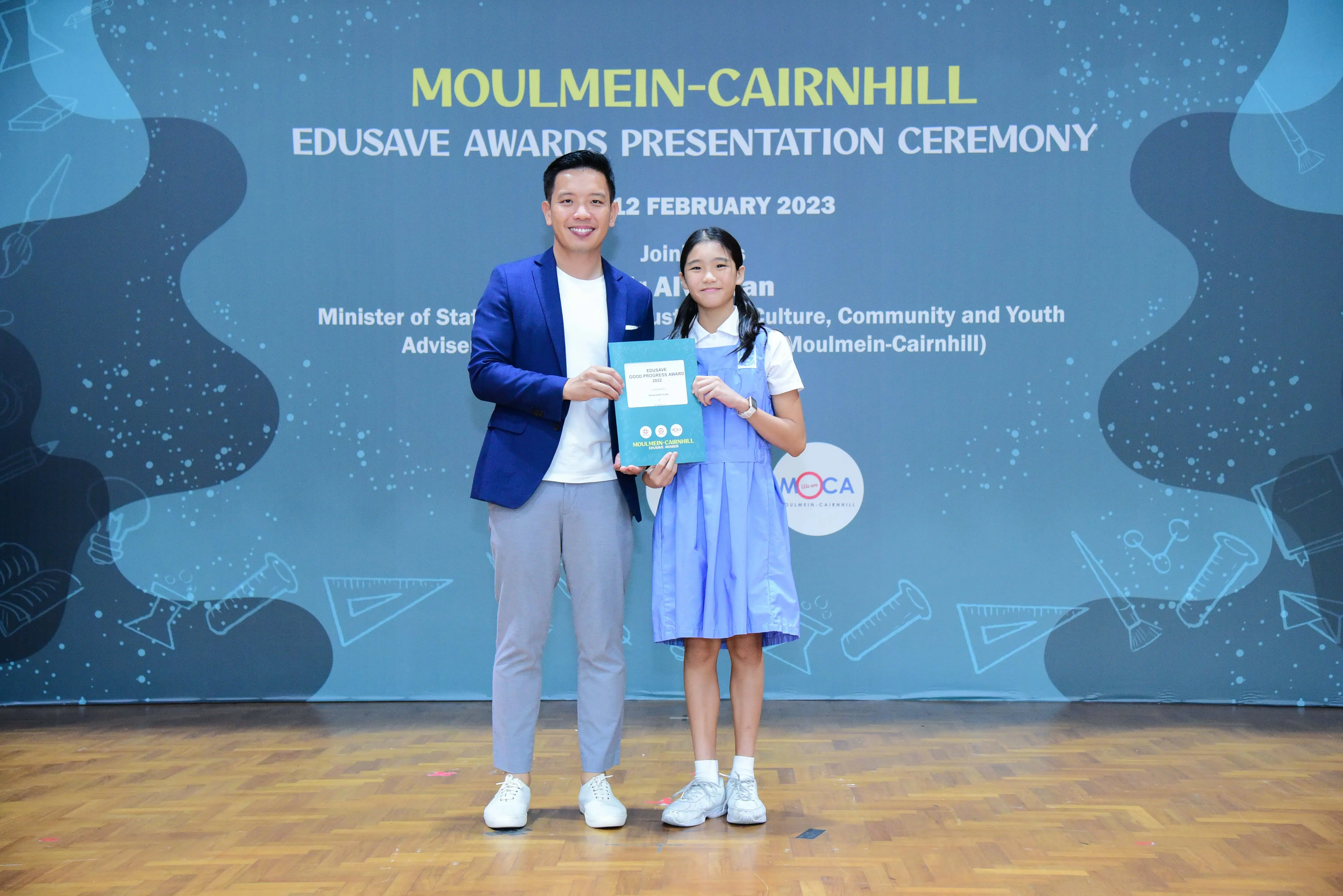 A man in a blue blazer and a girl in a school uniform hold an award at the Moulmein-Cairnhill Edusave Awards Ceremony.