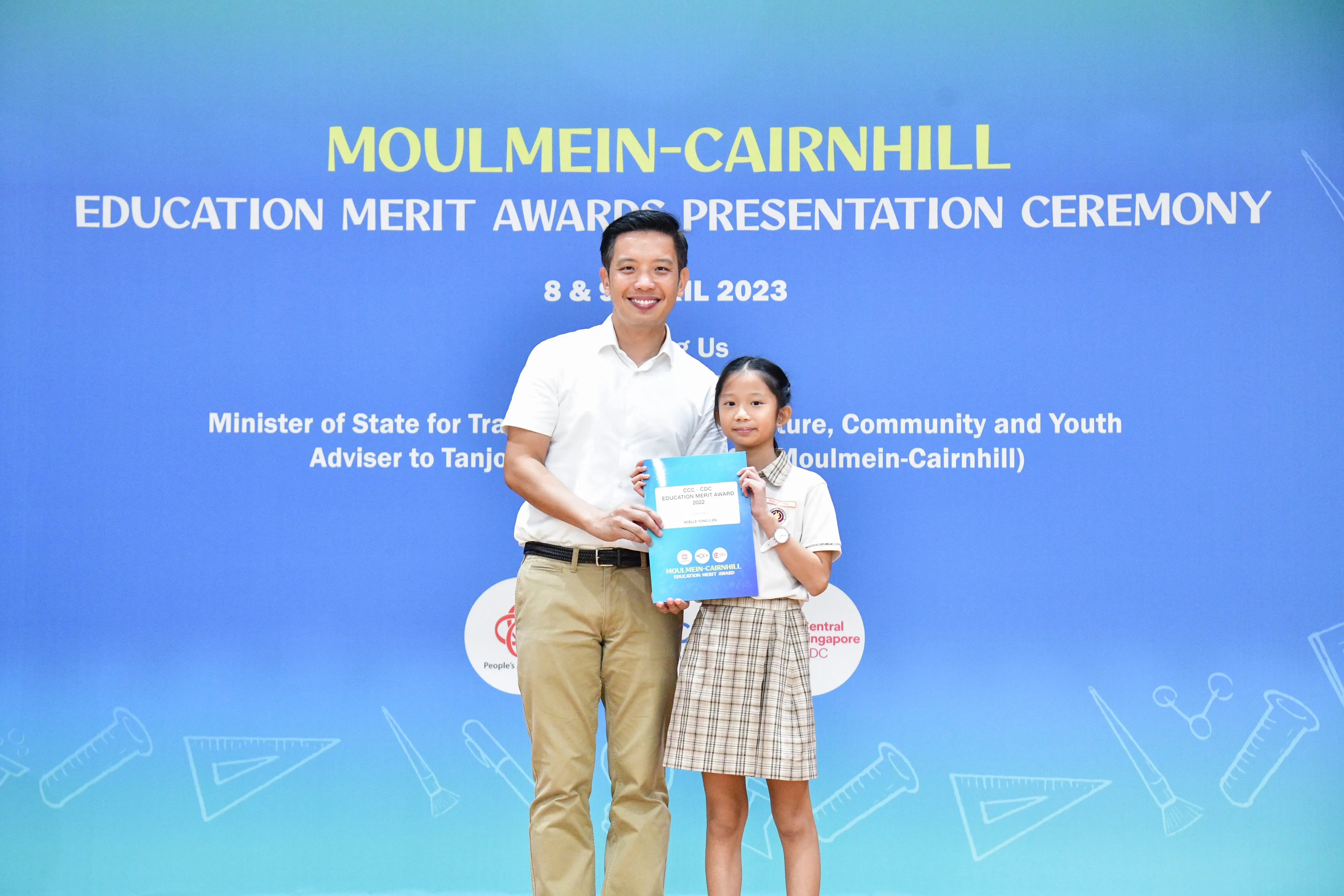 A man and a young girl hold a certificate in front of a Moulmein-Cairnhill Education Merit Awards 2023 banner.