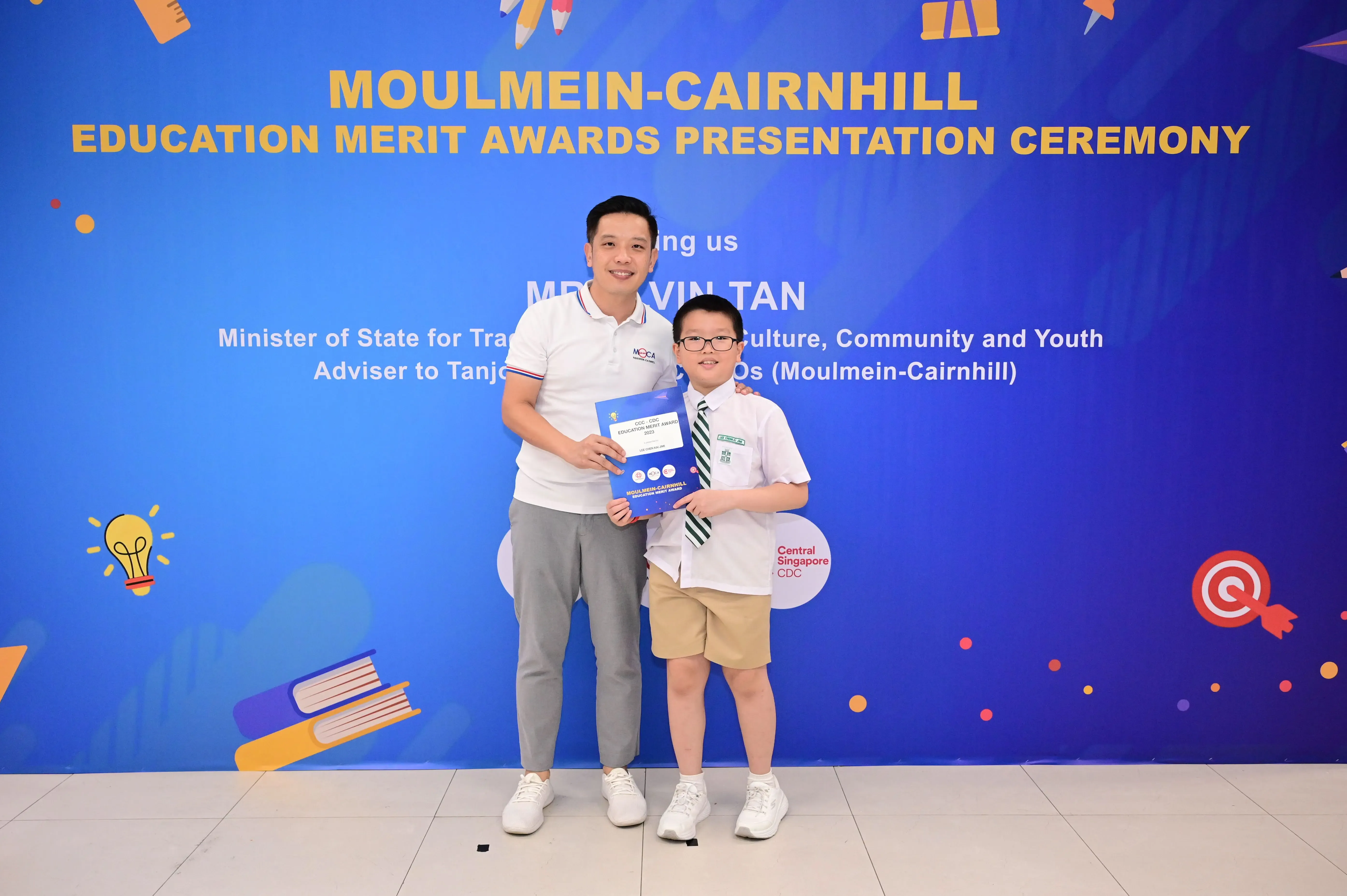 A man and a young boy in school uniform hold a certificate at the Moulmein-Cairnhill Education Merit Awards ceremony.