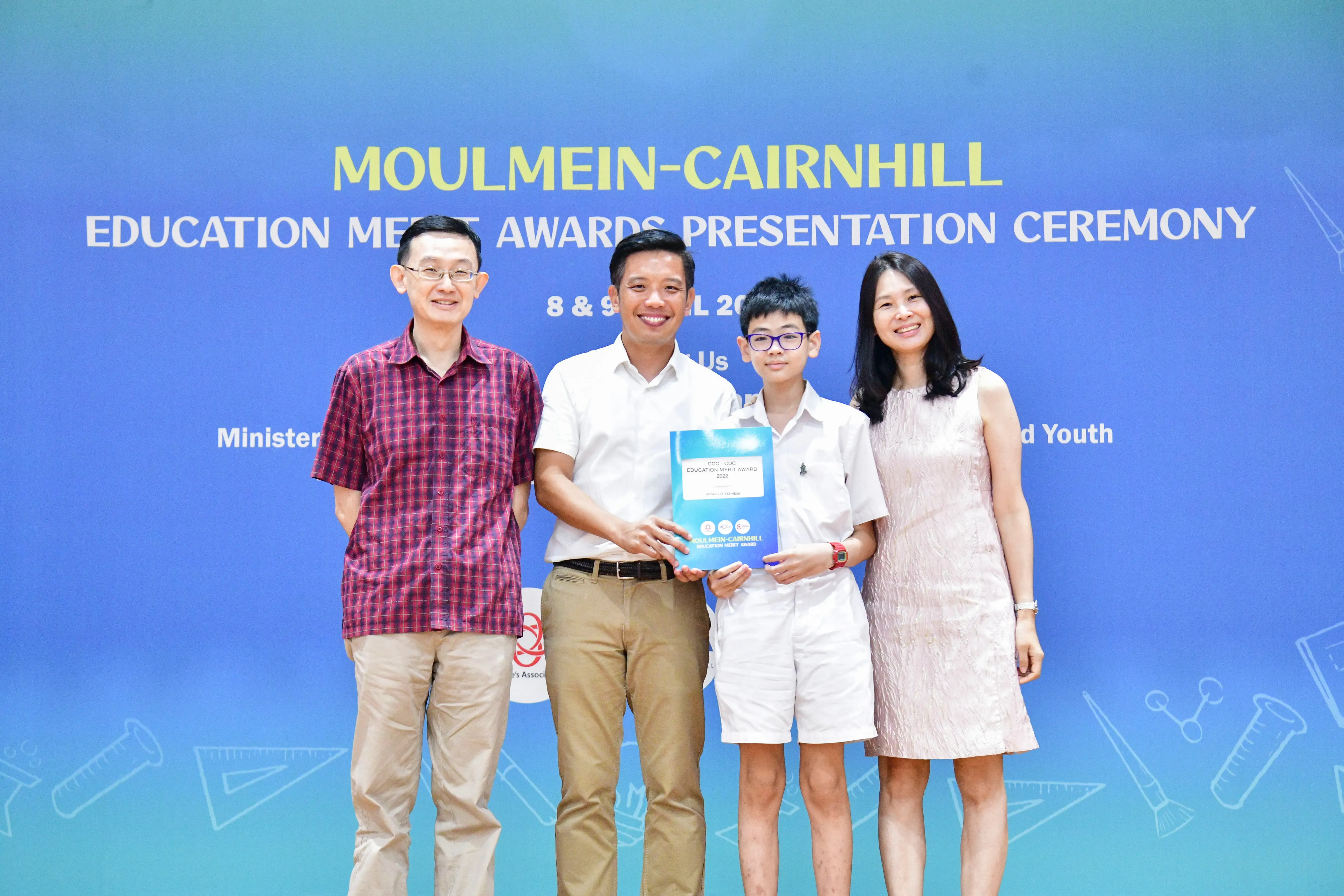 Four people pose at the Moulmein-Cairnhill Education Merit Awards Presentation Ceremony, holding a blue award certificate.