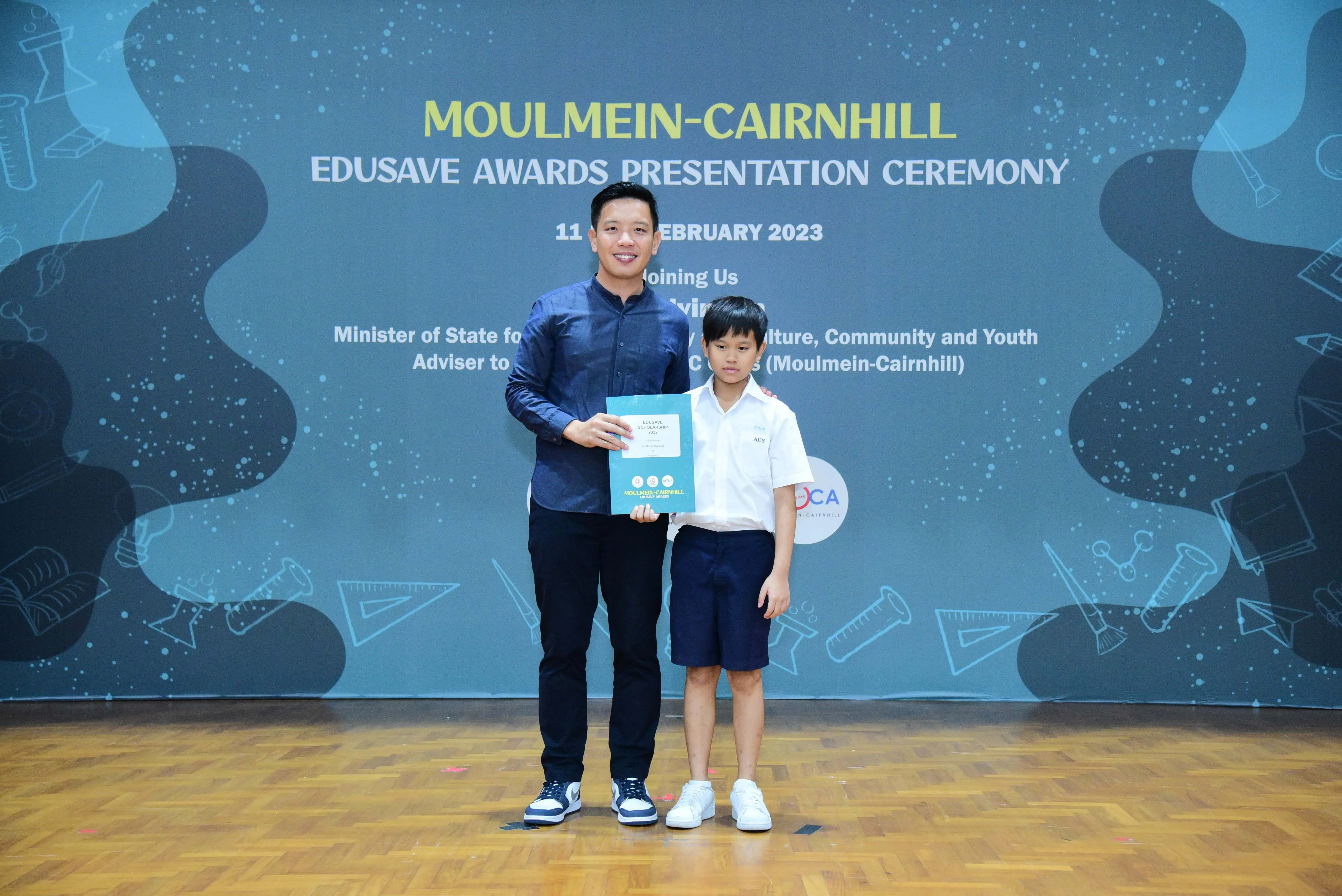 Adult man and young boy pose together holding a certificate at the Moulmein-Cairnhill Edusave Awards Ceremony 2023.