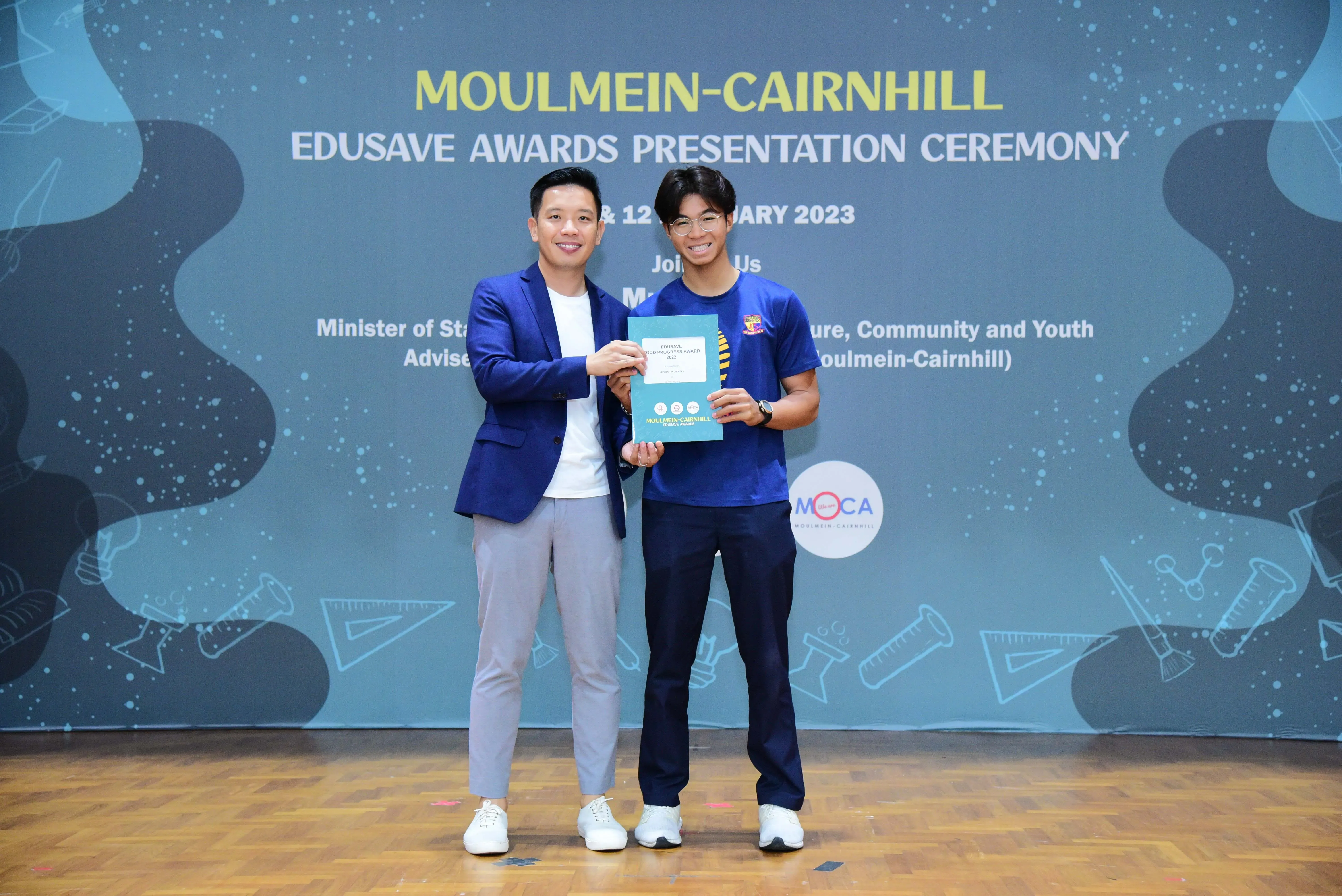 Two smiling men hold a certificate together at the Moulmein-Cairnhill Edusave Awards Presentation Ceremony 2023.