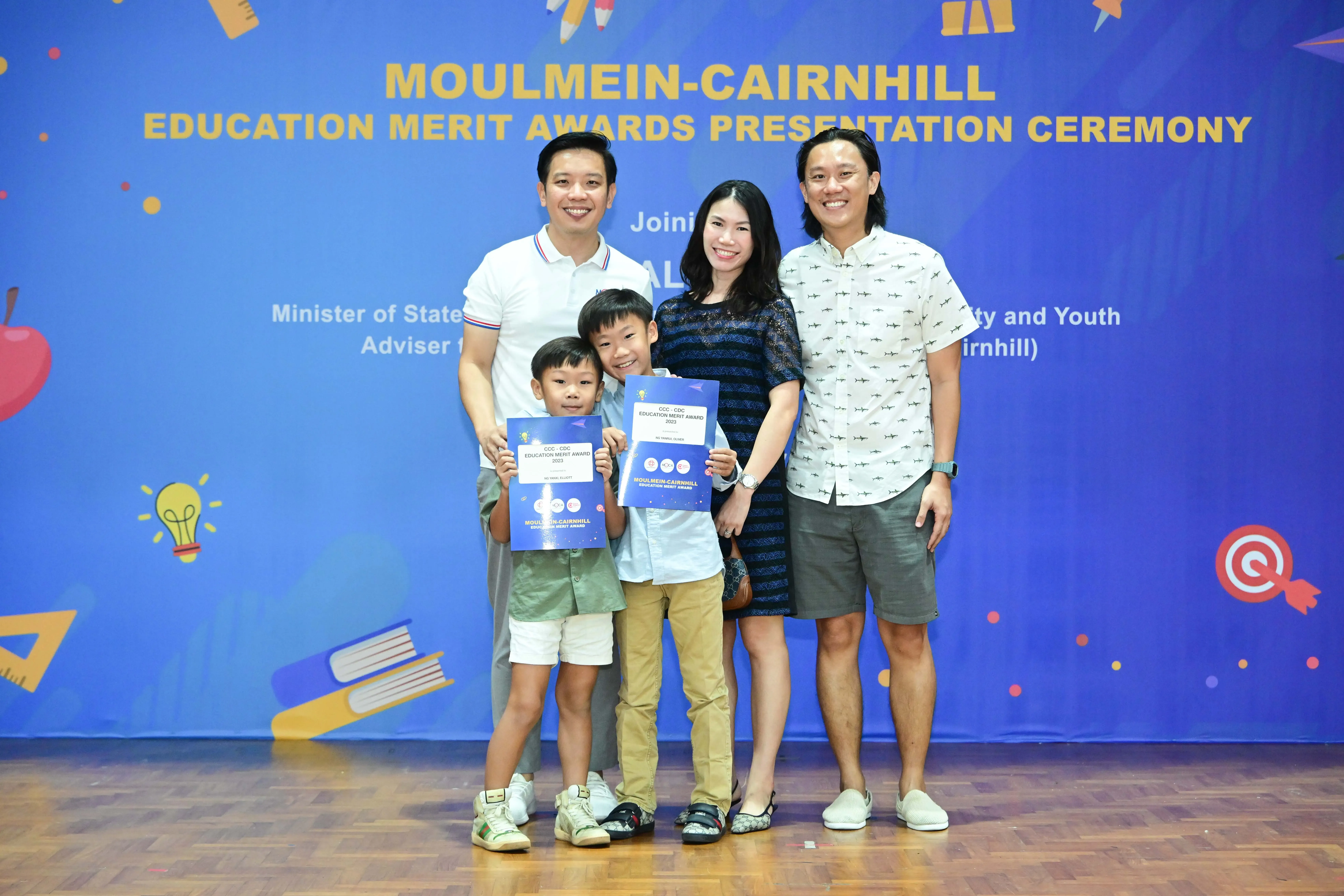 Five people smile in front of a Moulmein-Cairnhill Education Merit Awards banner, two children holding award certificates.