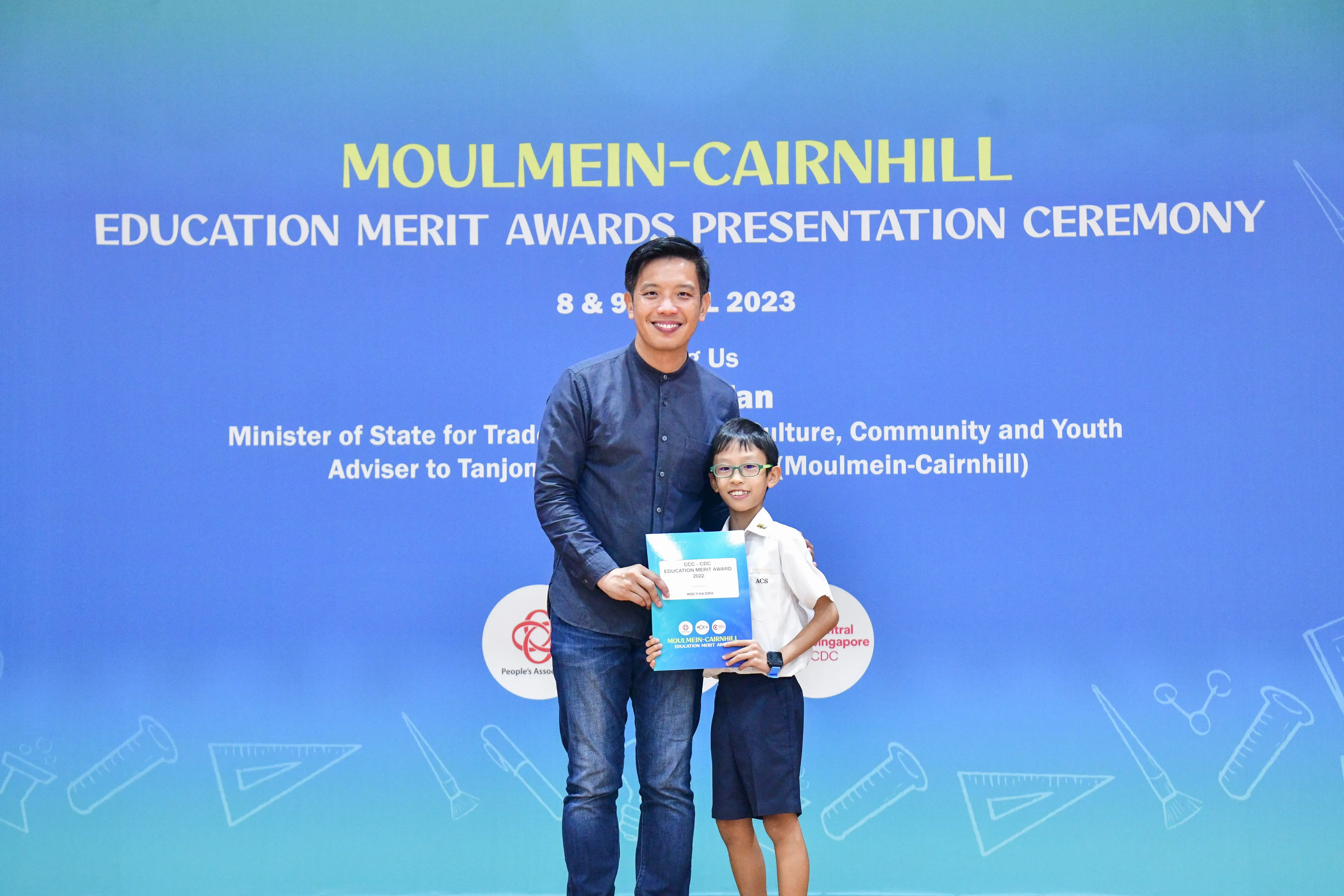 A man and a young boy holding a certificate stand before a Moulmein-Cairnhill Education Merit Awards banner dated 2023.
