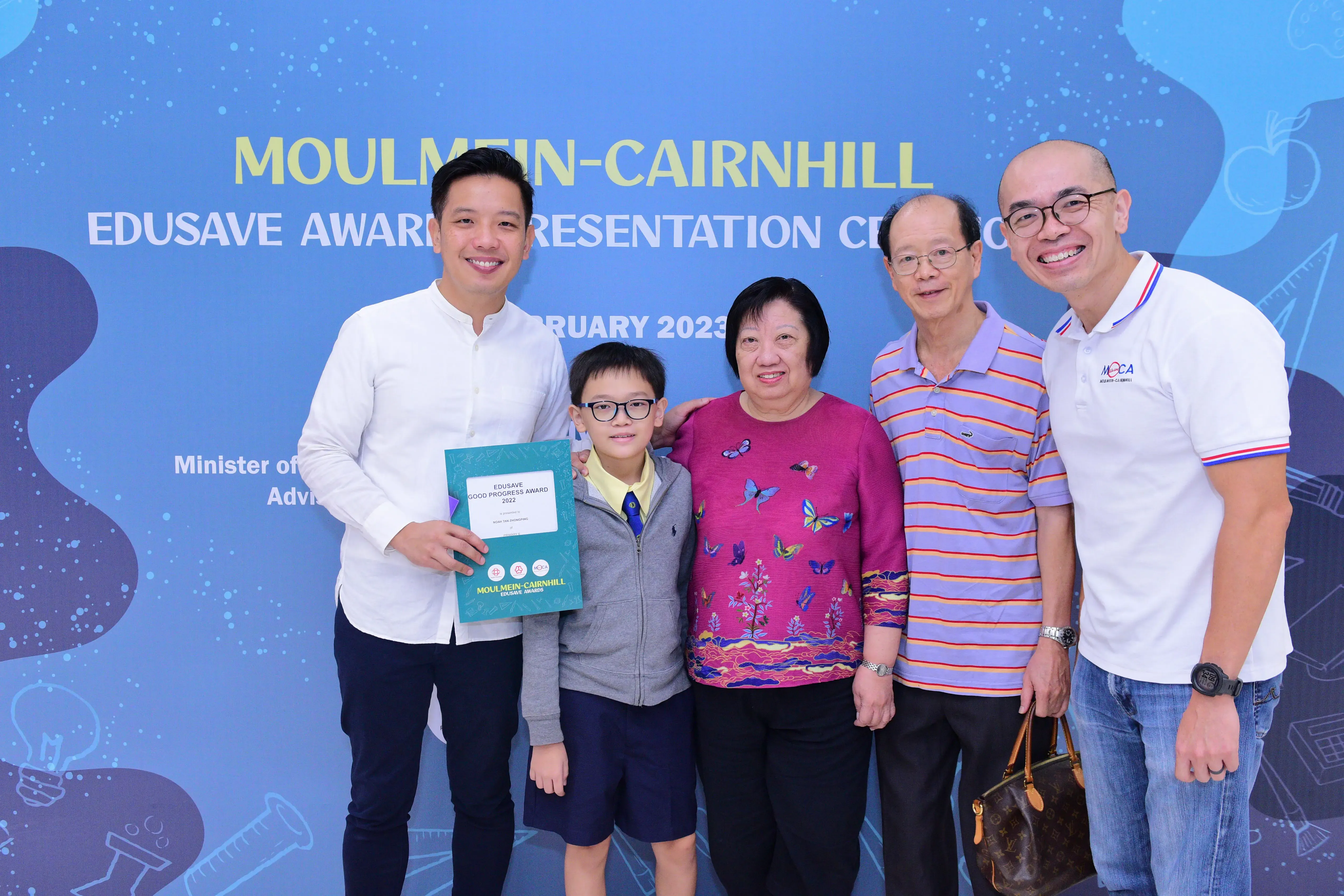Five people smile in front of a Moulmein-Cairnhill Edusave Award Presentation Ceremony 2023 banner, boy holding a certificate.