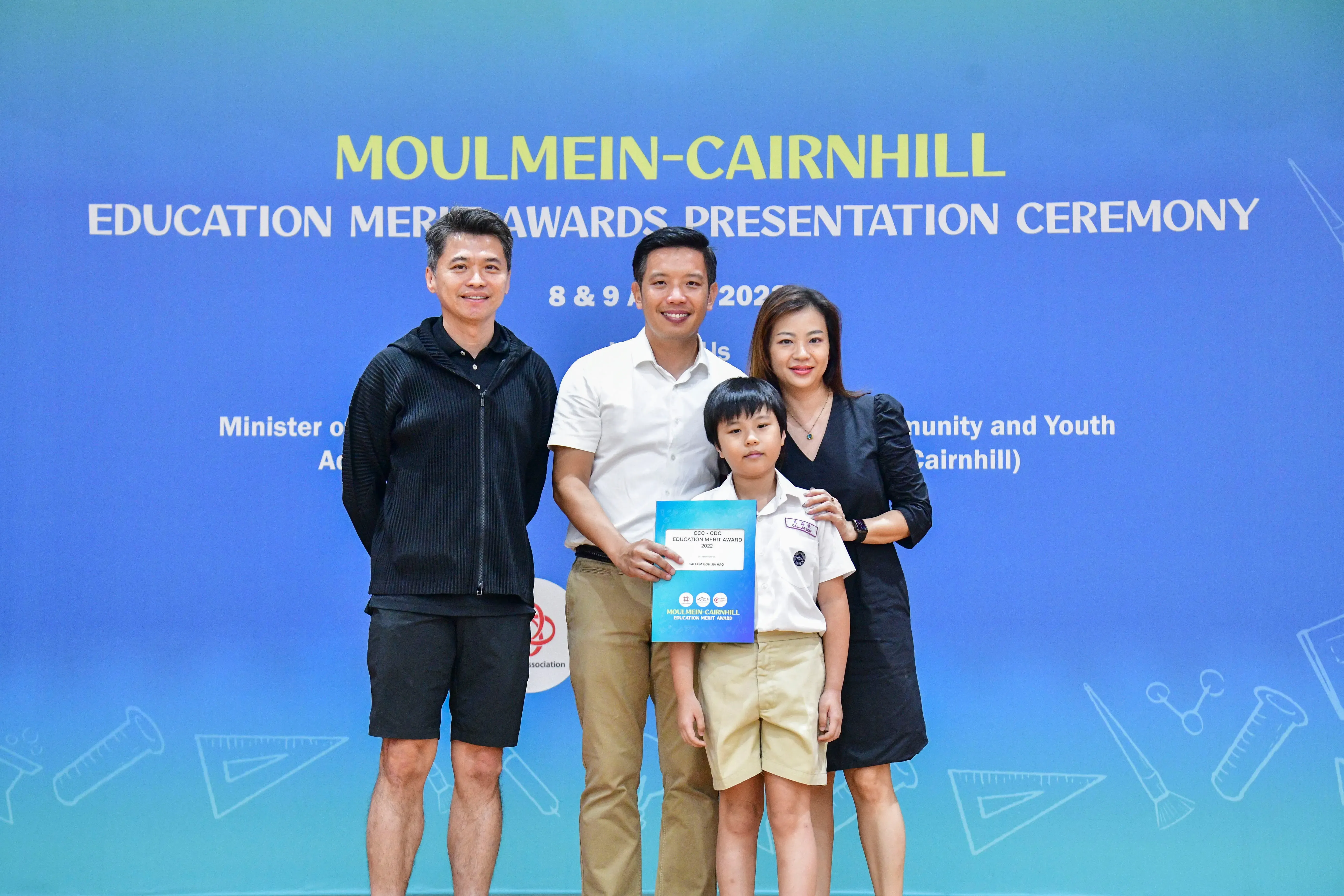 Three adults and a child pose before a Moulmein-Cairnhill Education Merit Awards Presentation Ceremony banner.