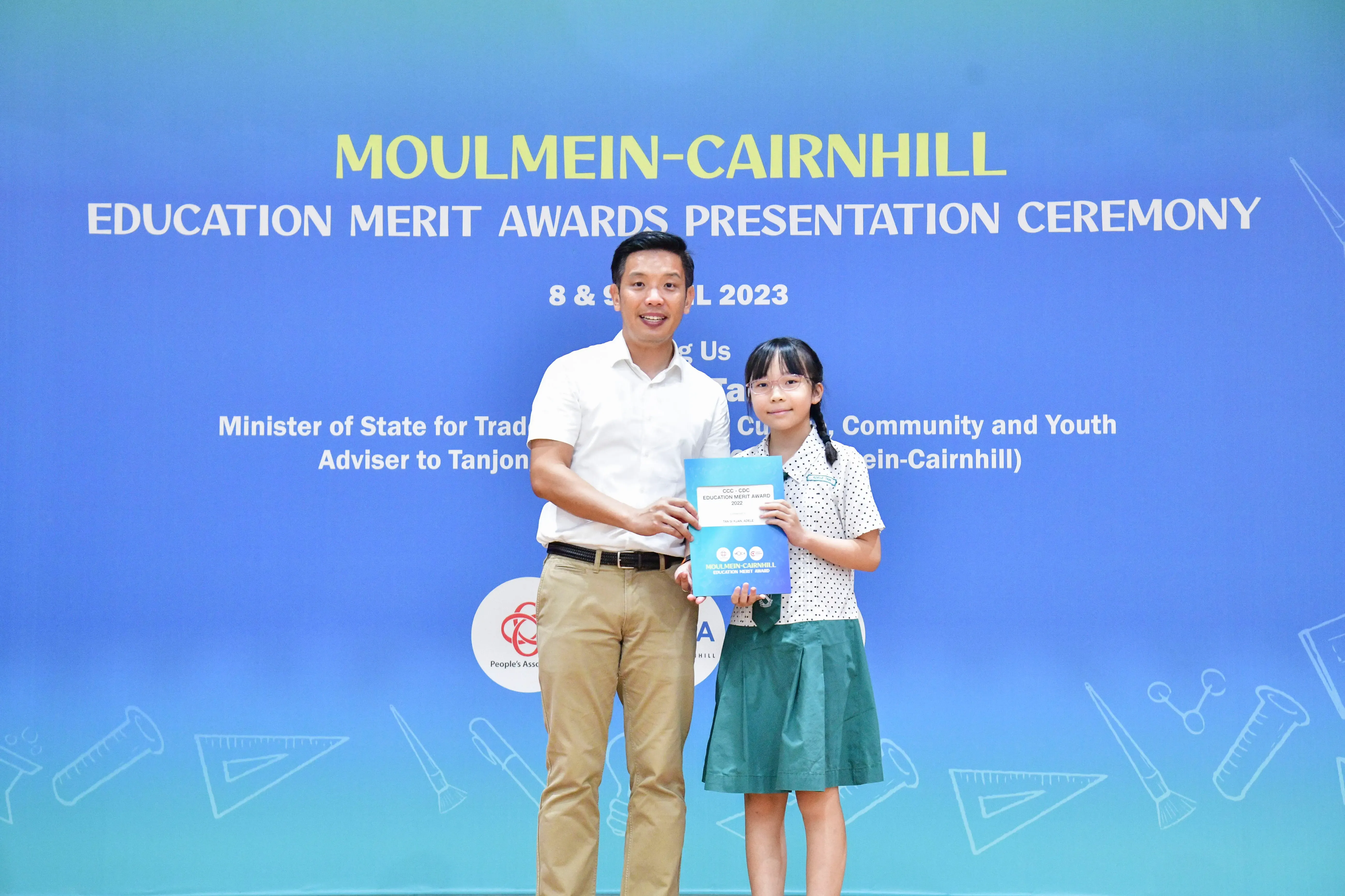 A man and a young girl hold a blue award folder at the Moulmein-Cairnhill Education Merit Awards Ceremony 2023.