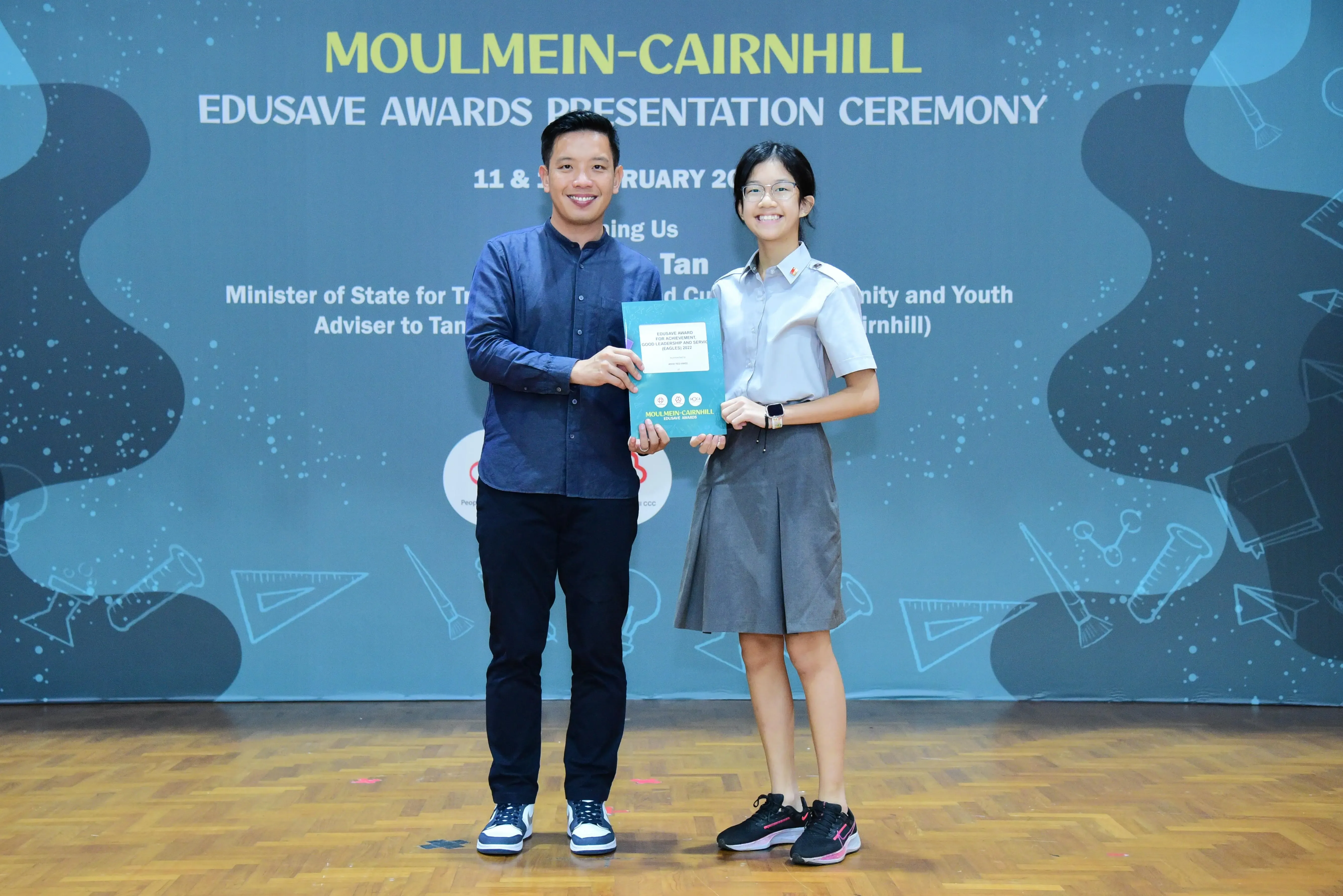 Two smiling people hold a teal award booklet at the Moulmein-Cairnhill Edusave Awards Presentation Ceremony.