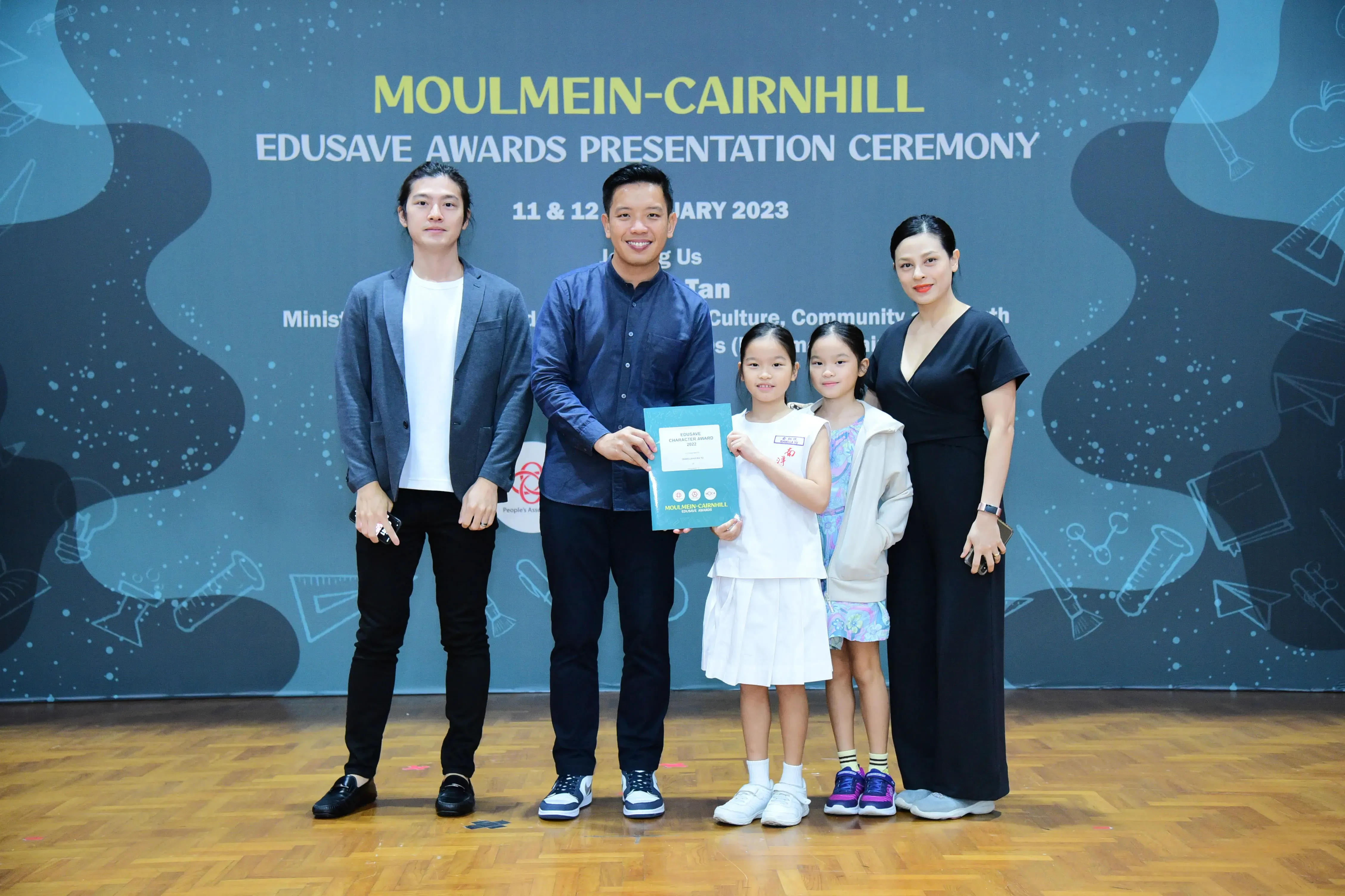 Five people pose at the Moulmein-Cairnhill Edusave Awards Presentation Ceremony 2023, with a man handing a certificate to two young girls.