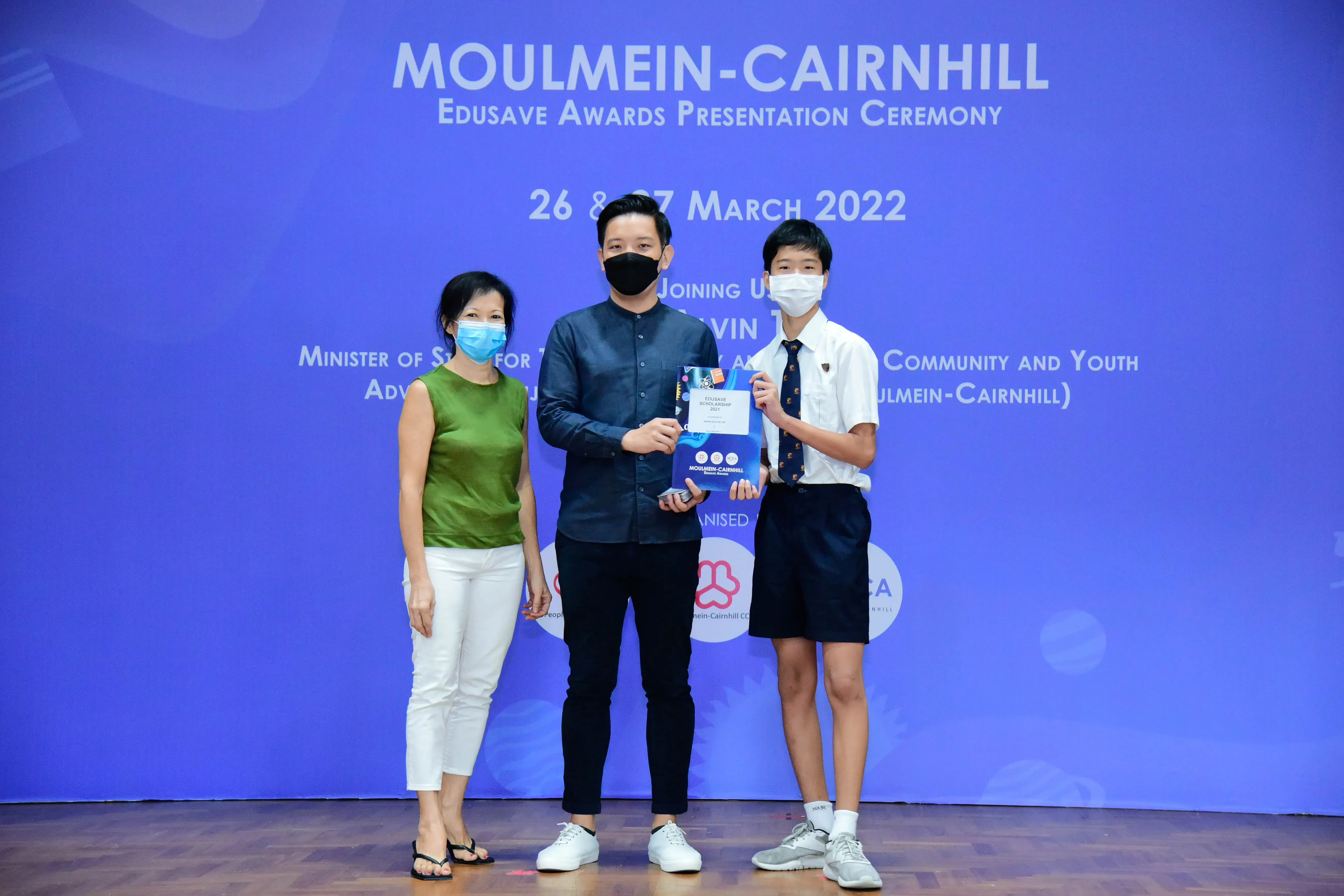 Three masked people stand before a blue Moulmein-Cairnhill Edusave Awards Presentation Ceremony backdrop, holding an award.