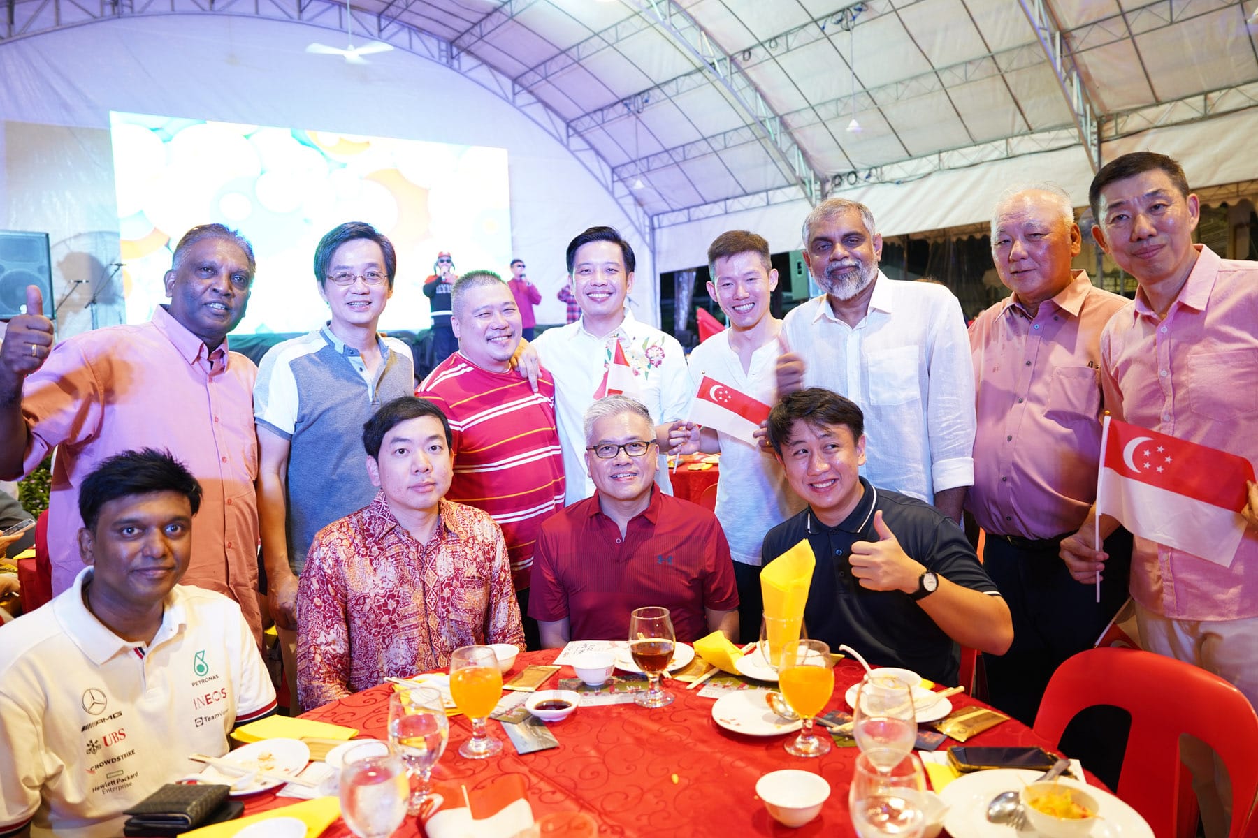 Group of men posing and smiling at a dinner event under a tent, holding Singapore flags near a red table with drinks.