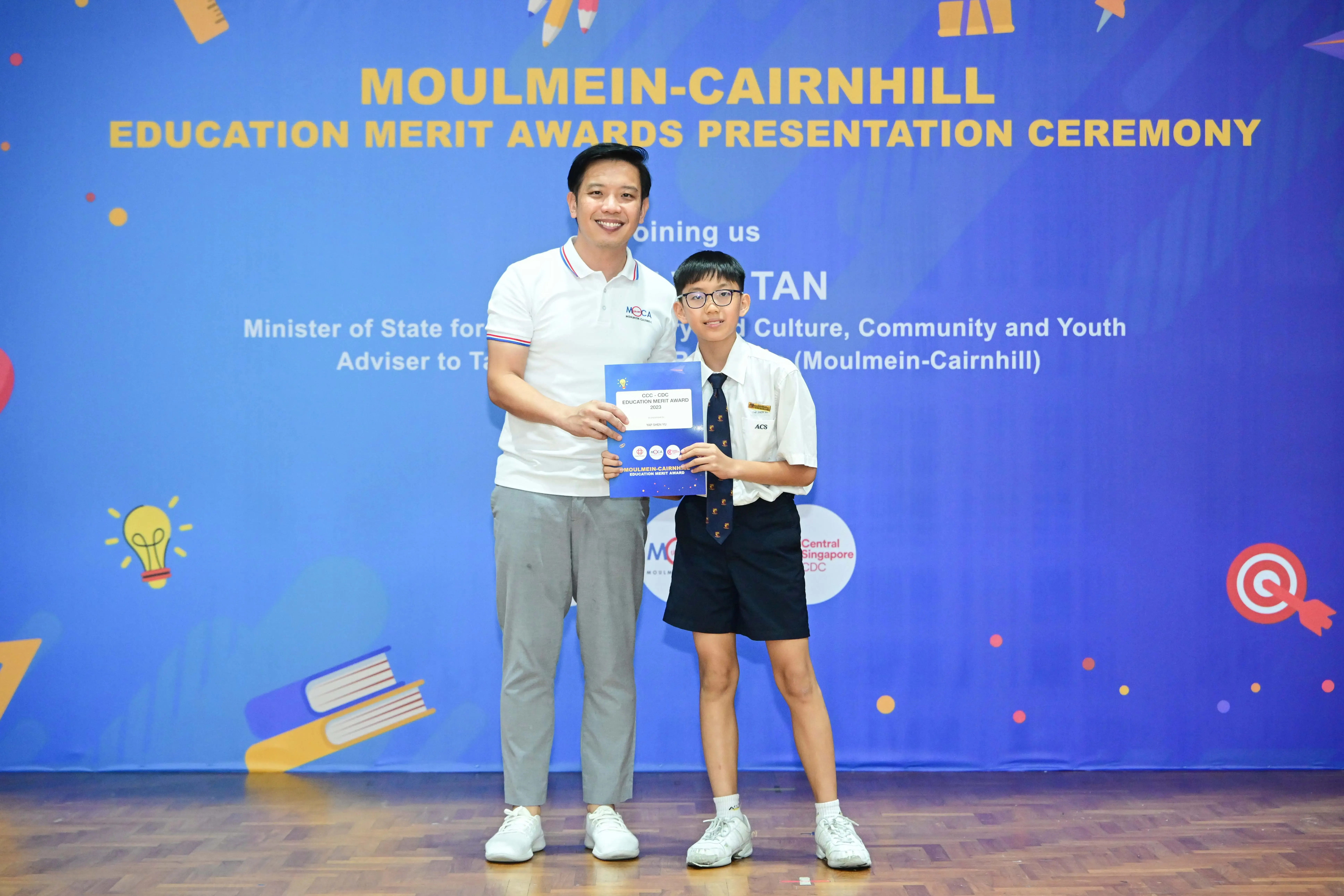 An adult and a young student hold an award certificate at the Moulmein-Cairnhill Education Merit Awards ceremony.