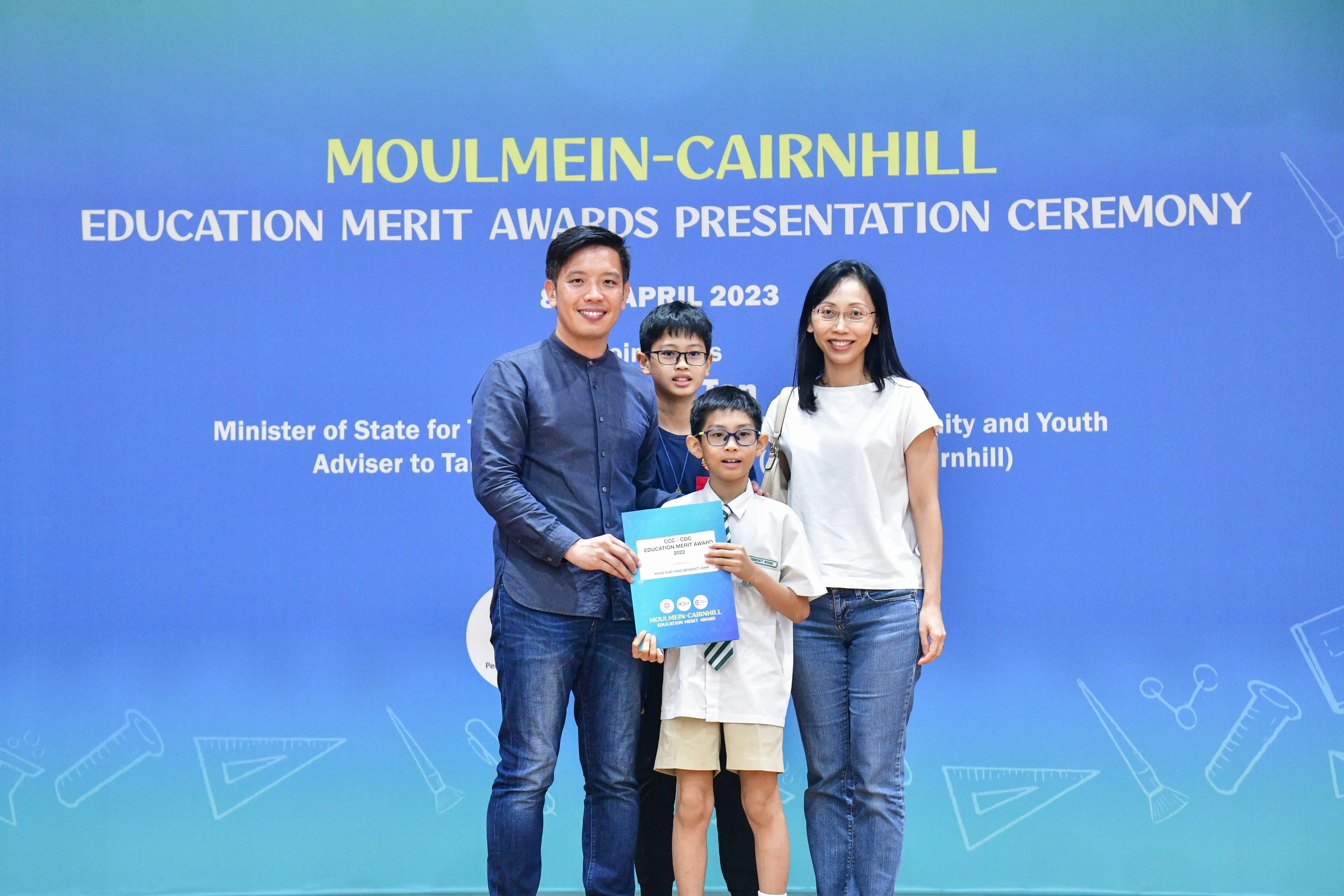 Family of four poses before a Moulmein-Cairnhill Education Merit Awards banner; a boy holds an award certificate.