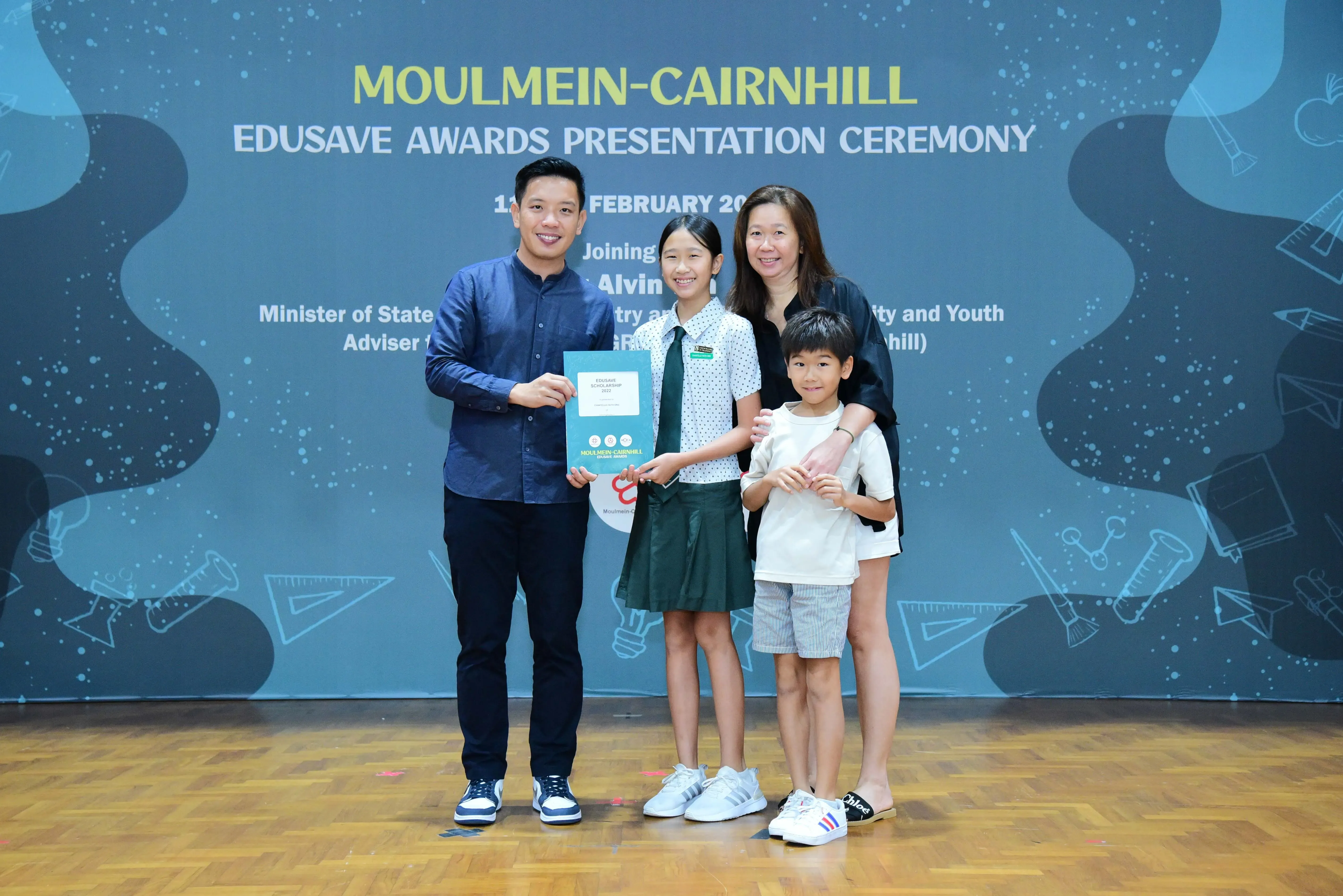 Four people pose on a stage at the Moulmein-Cairnhill Edusave Awards Presentation Ceremony holding a certificate.