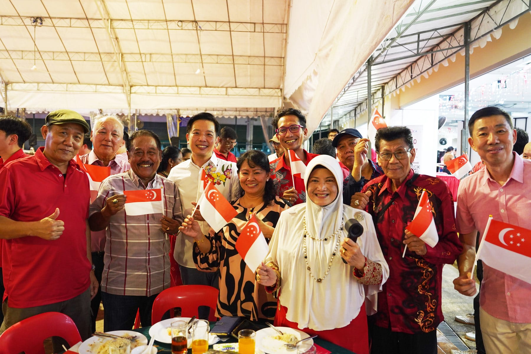 A smiling group of people holding small Singapore flags gathered under a tent at an outdoor community event.