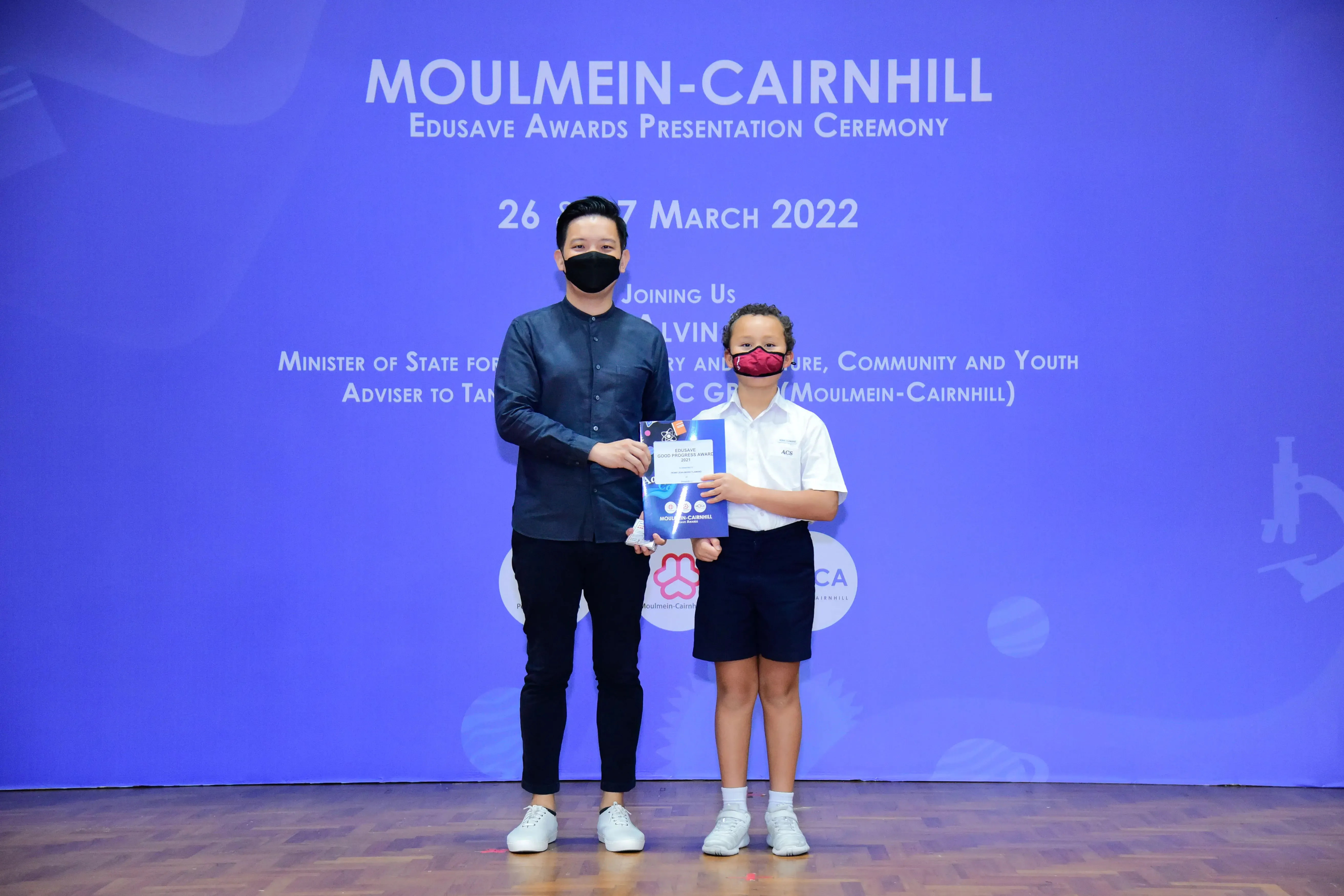 An adult and a child in masks hold a certificate on stage at the Moulmein-Cairnhill Edusave Awards Ceremony 2022.