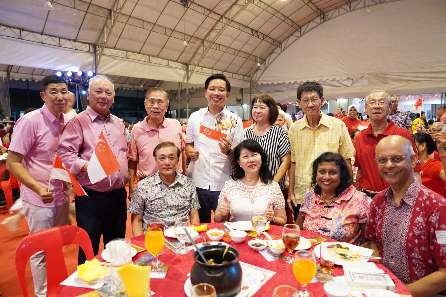 A diverse group of smiling people pose at a red-draped dinner table under a tent, holding small Singapore flags.