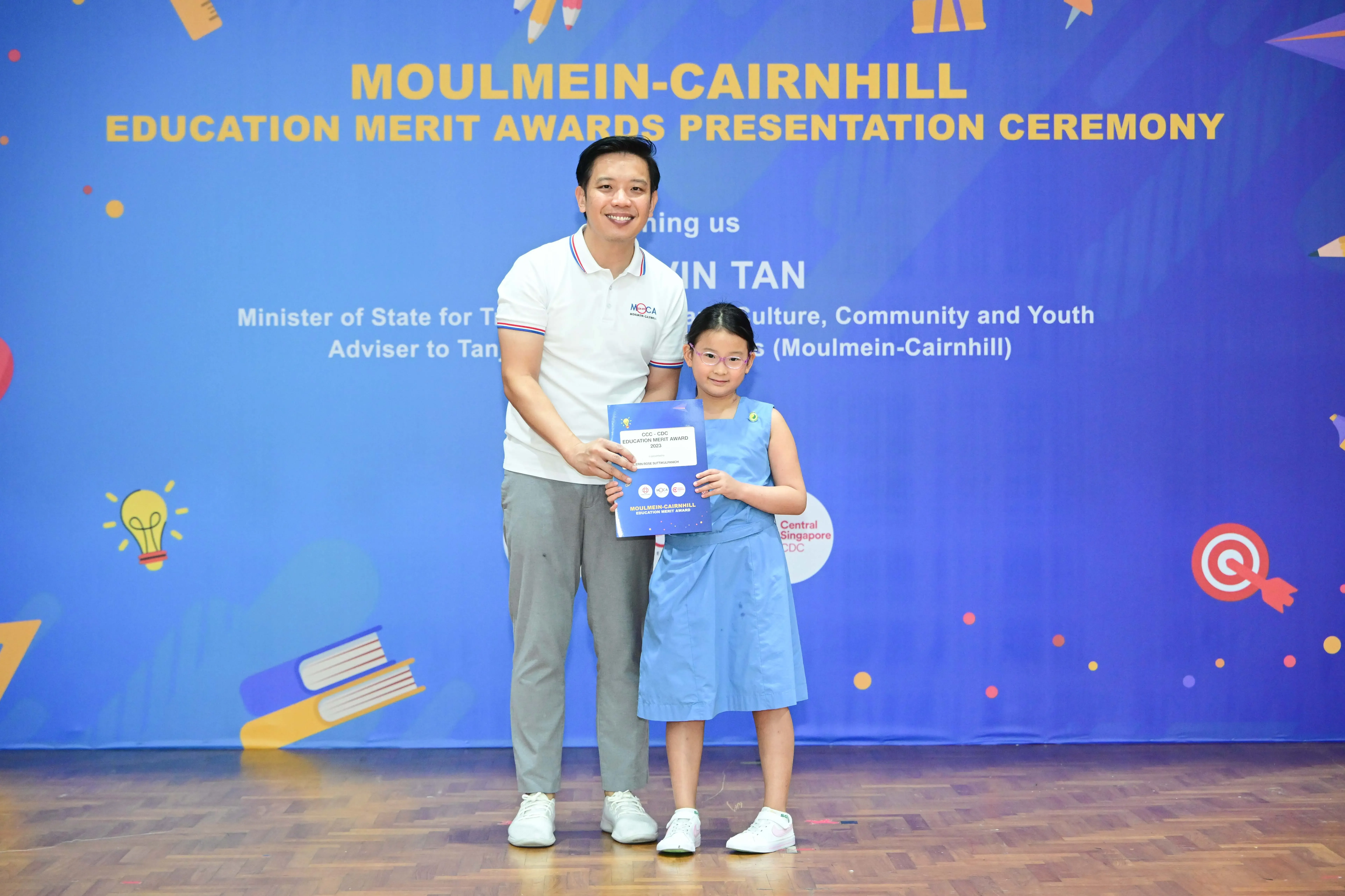 A man and a young girl hold a certificate in front of a Moulmein-Cairnhill Education Merit Awards banner.
