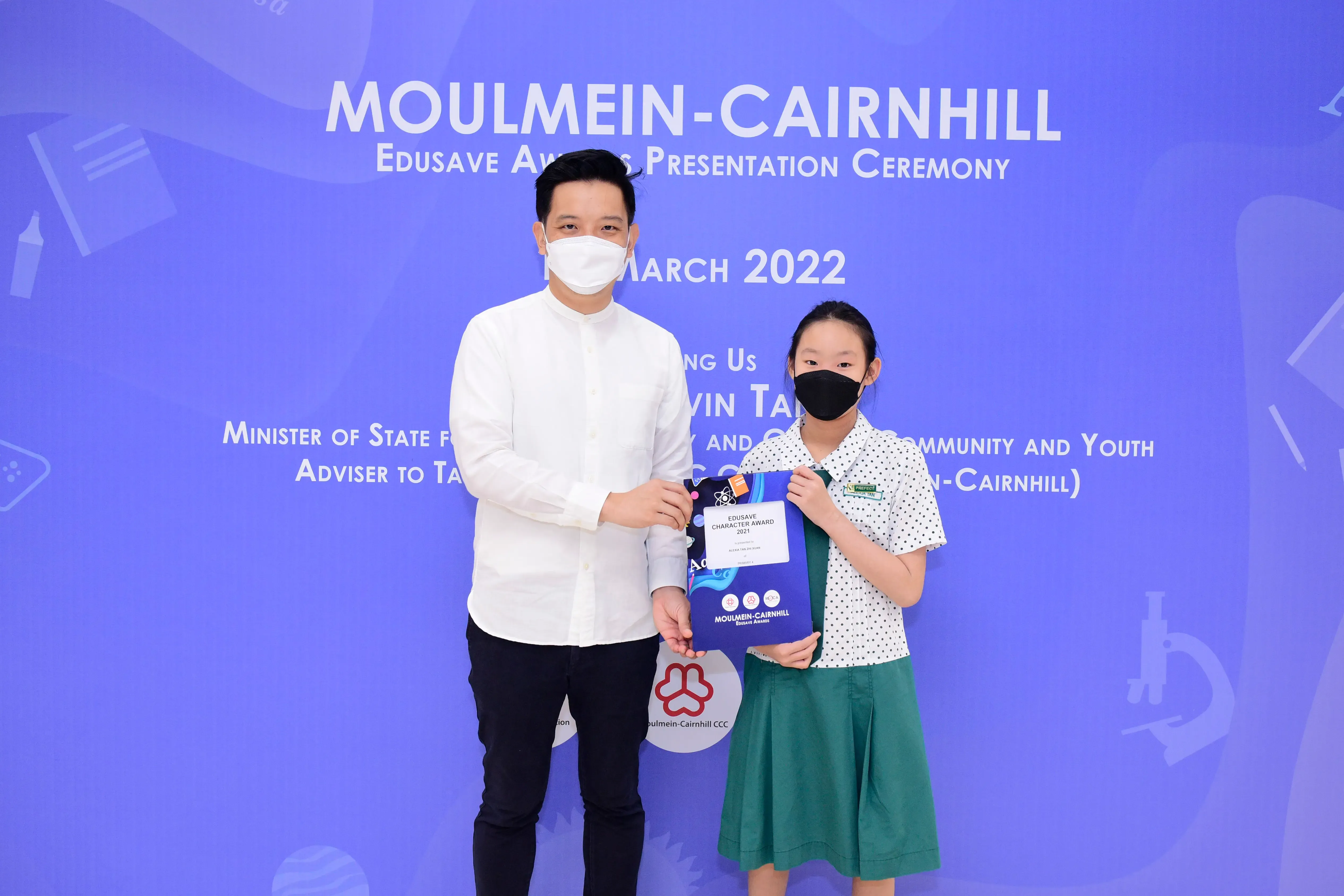 An adult and a young girl in masks hold an Edusave Character Award at the Moulmein-Cairnhill ceremony, March 2022.