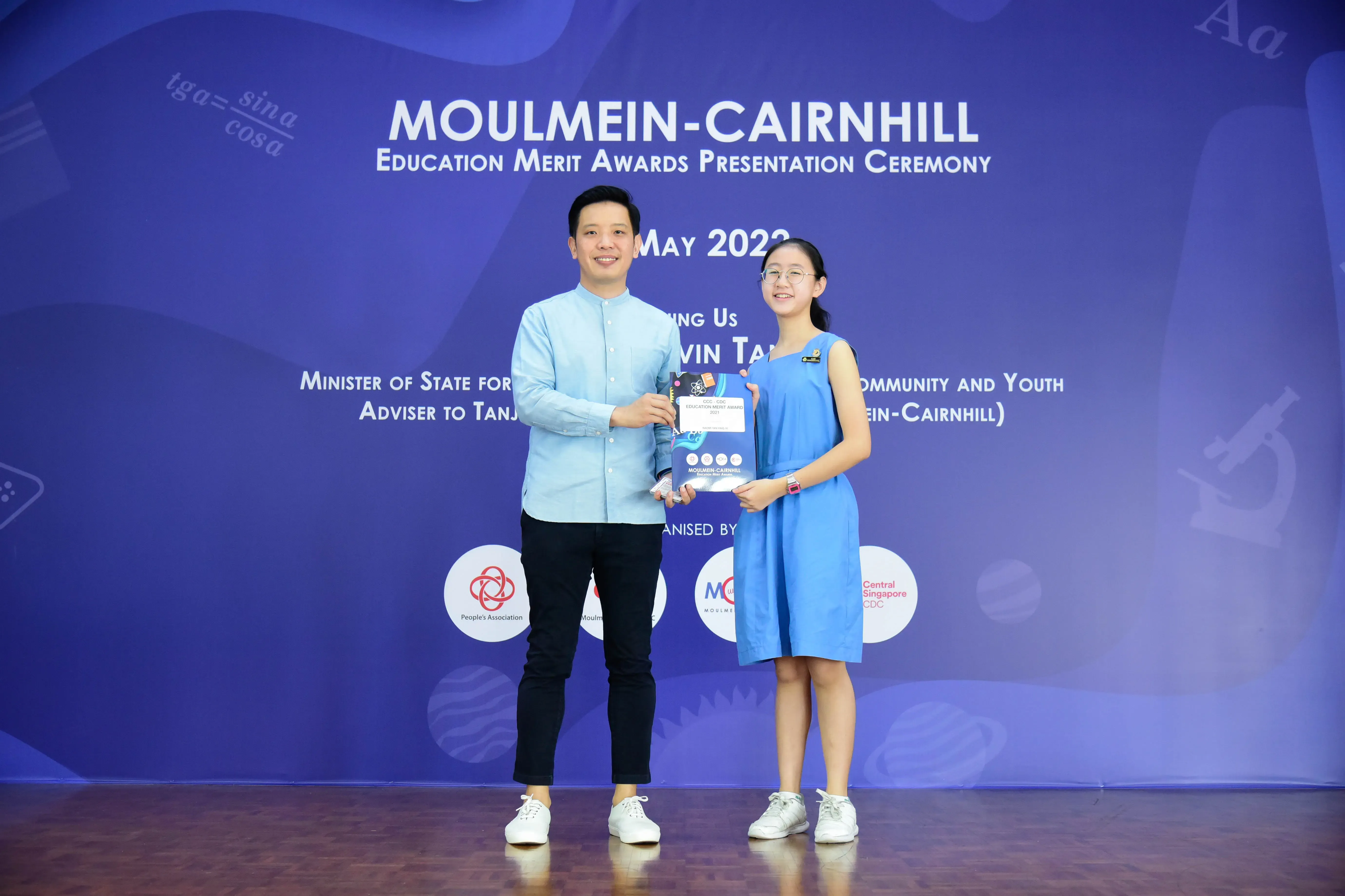 A man and a young girl hold an award together on stage at the Moulmein-Cairnhill Education Merit Awards Ceremony 2022.