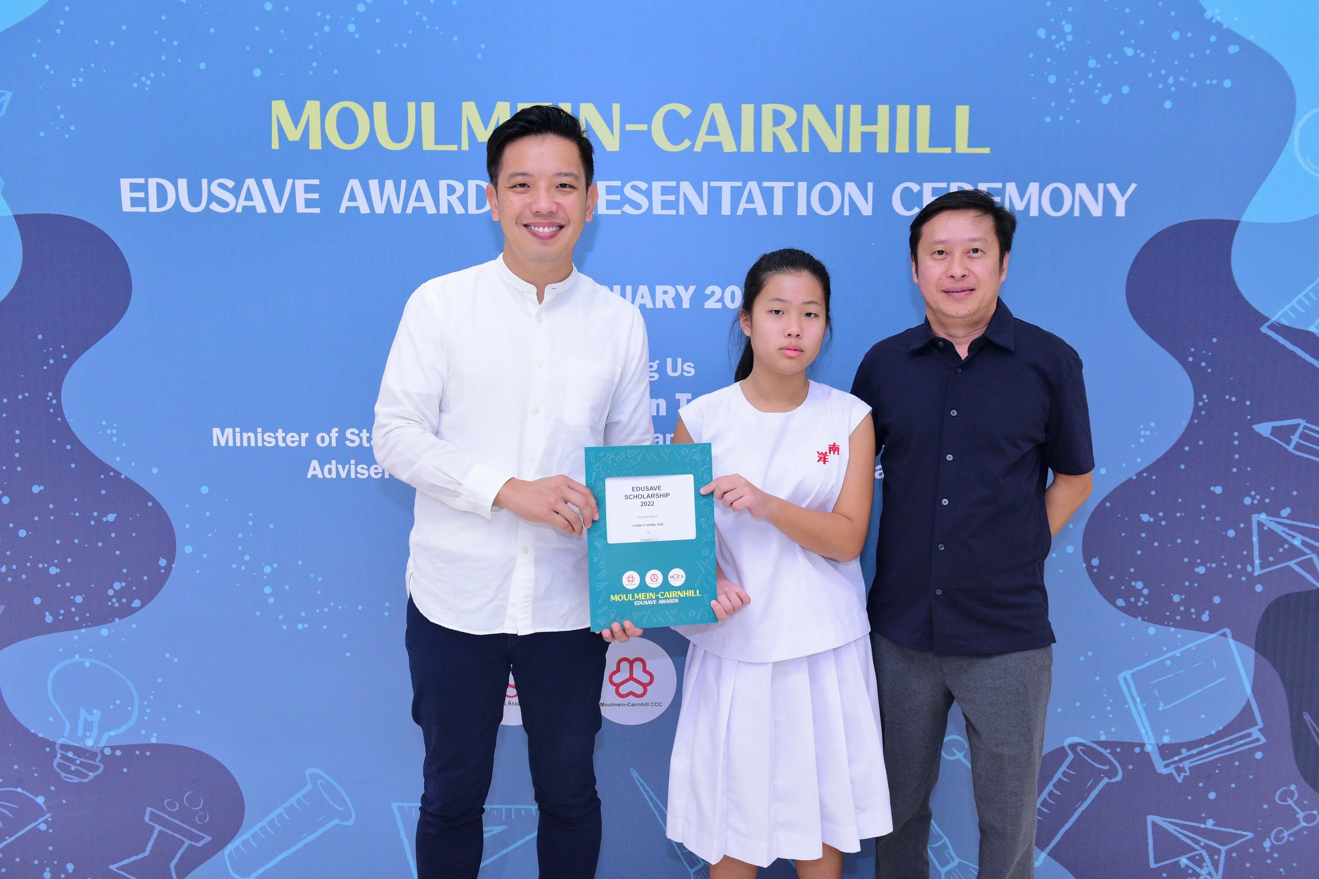 Three people pose at the Moulmein-Cairnhill Edusave Awards ceremony, with a girl holding a teal scholarship certificate.