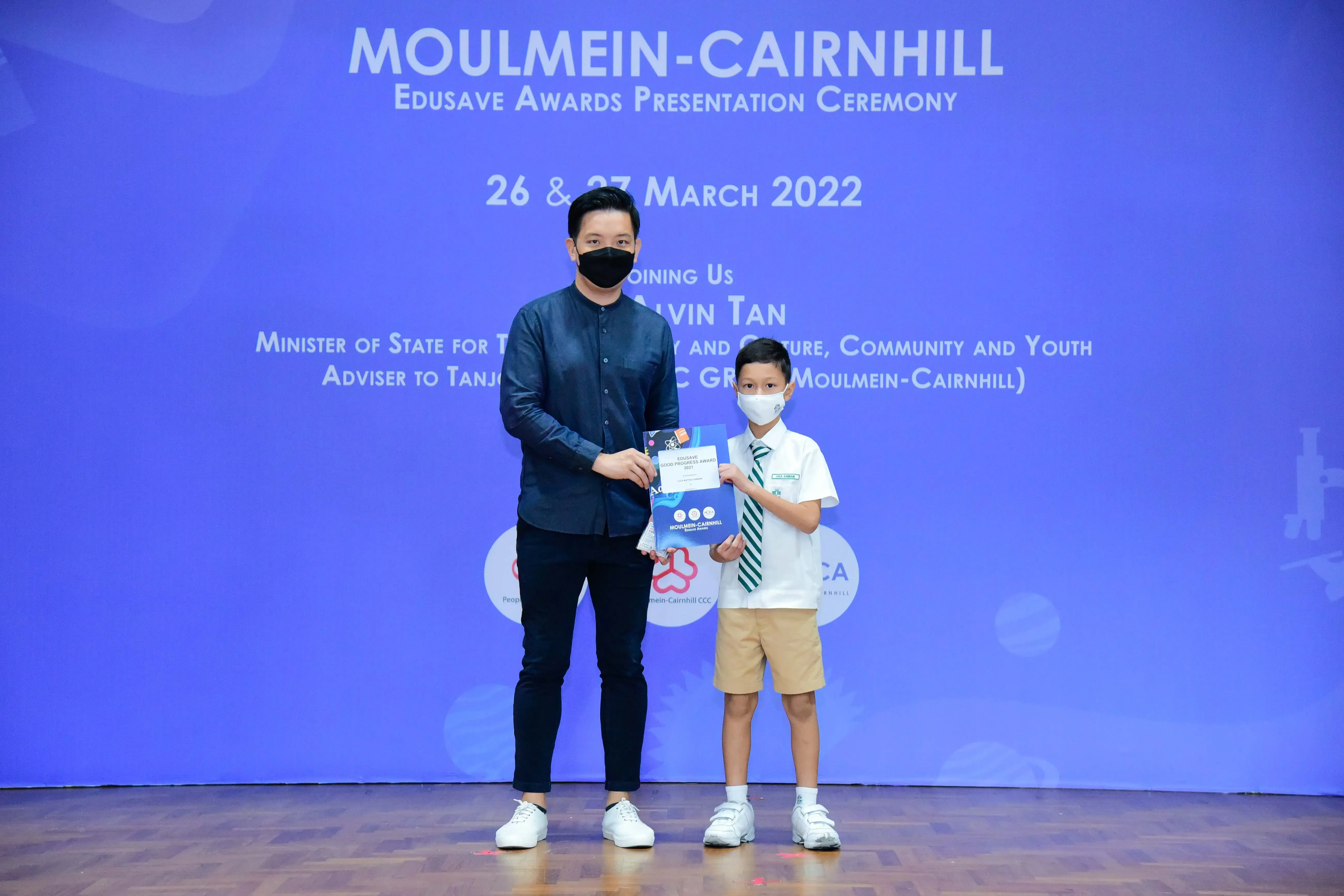 Adult and young student in masks hold an award at Moulmein-Cairnhill Edusave Awards Ceremony, March 2022.