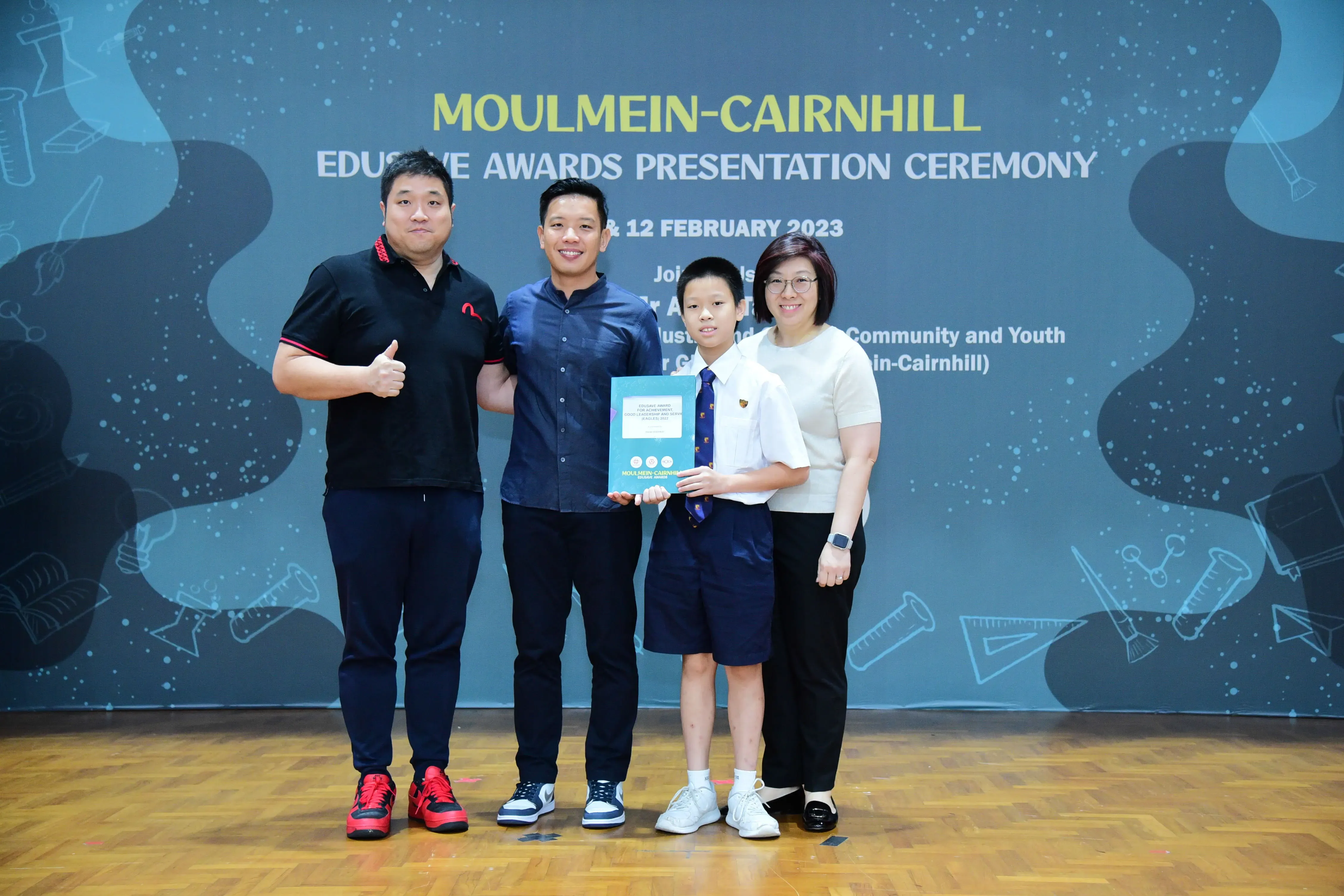 Three adults and a young boy in school uniform hold an award at the Moulmein-Cairnhill Edusave Awards Ceremony 2023.