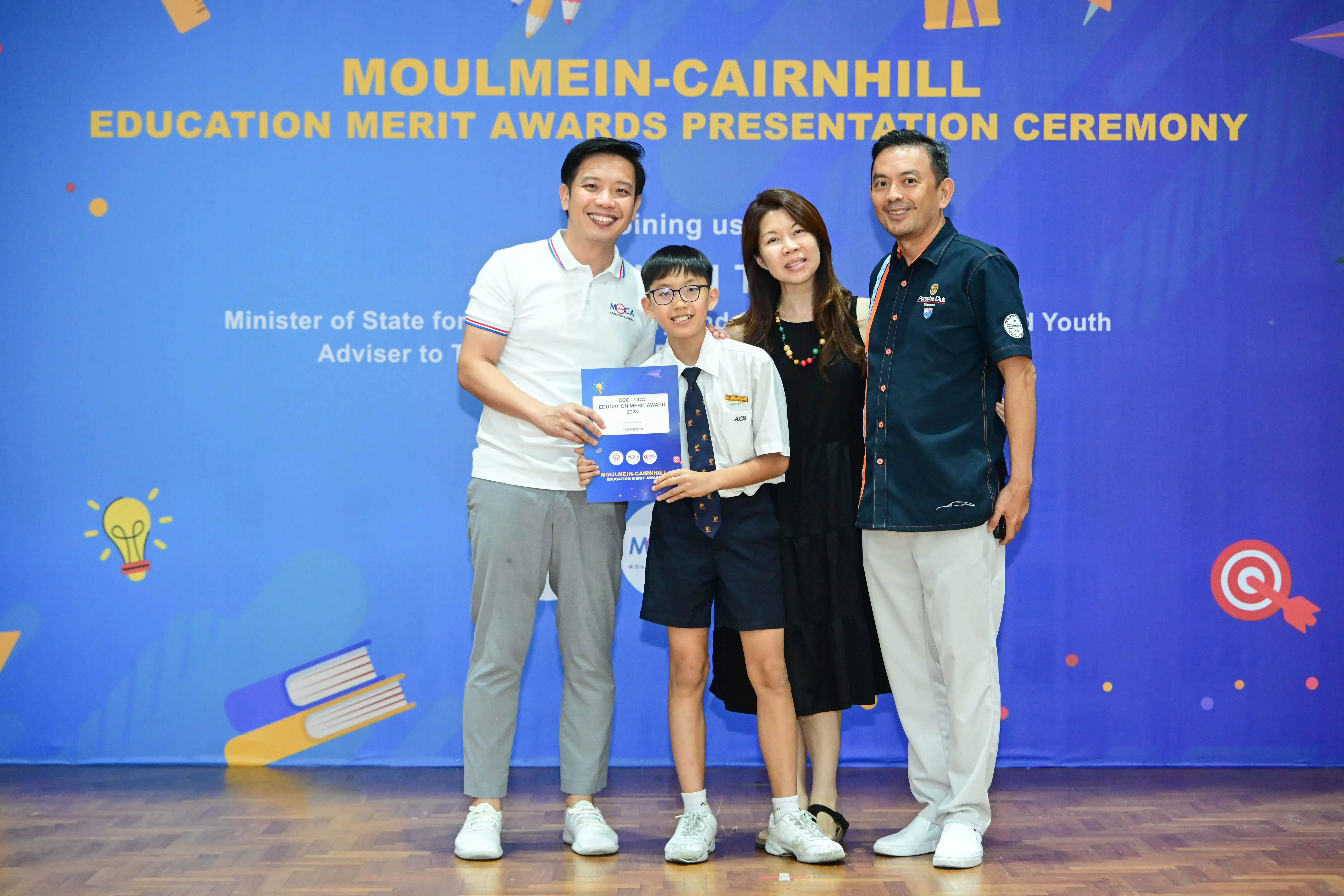 A boy in school uniform holds an award certificate with three adults at the Moulmein-Cairnhill Education Merit Awards ceremony.