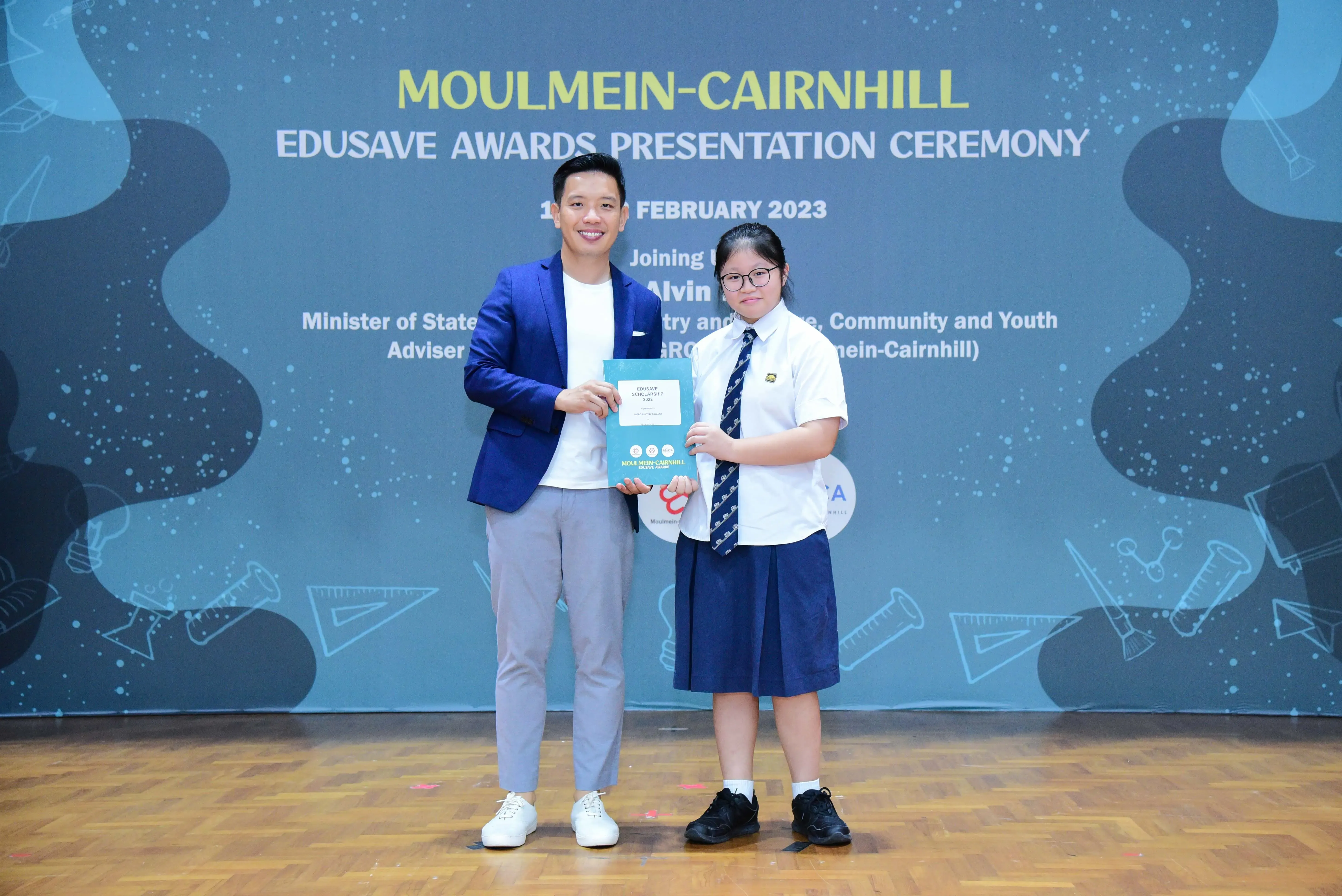 A man in a blue blazer presents a certificate to a uniformed student at the Moulmein-Cairnhill Edusave Awards Ceremony 2023.