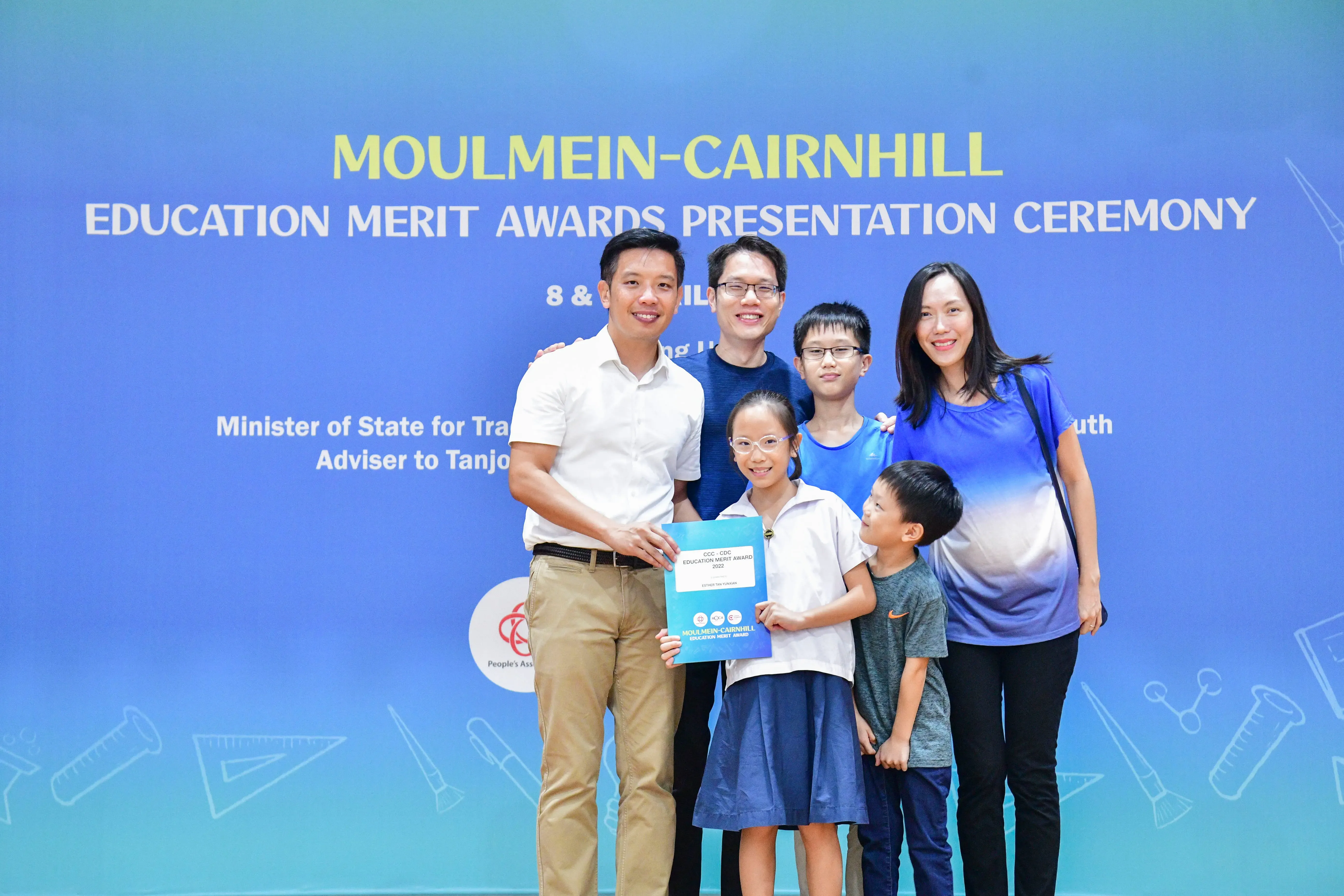 A girl holds an award certificate with a family and an official at the Moulmein-Cairnhill Education Merit Awards ceremony.