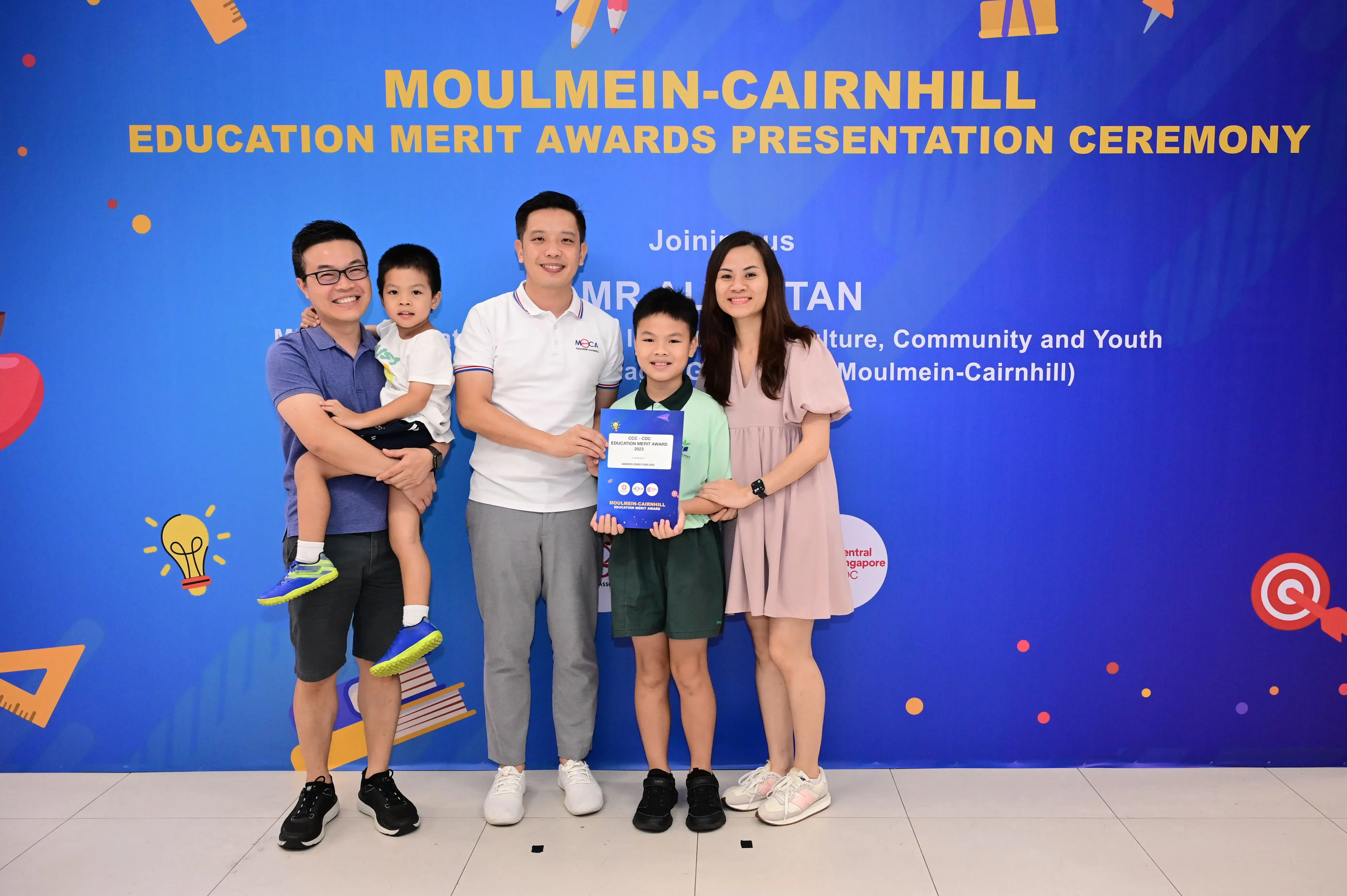 Five people pose before a Moulmein-Cairnhill Education Merit Awards banner, with a boy holding a blue certificate.