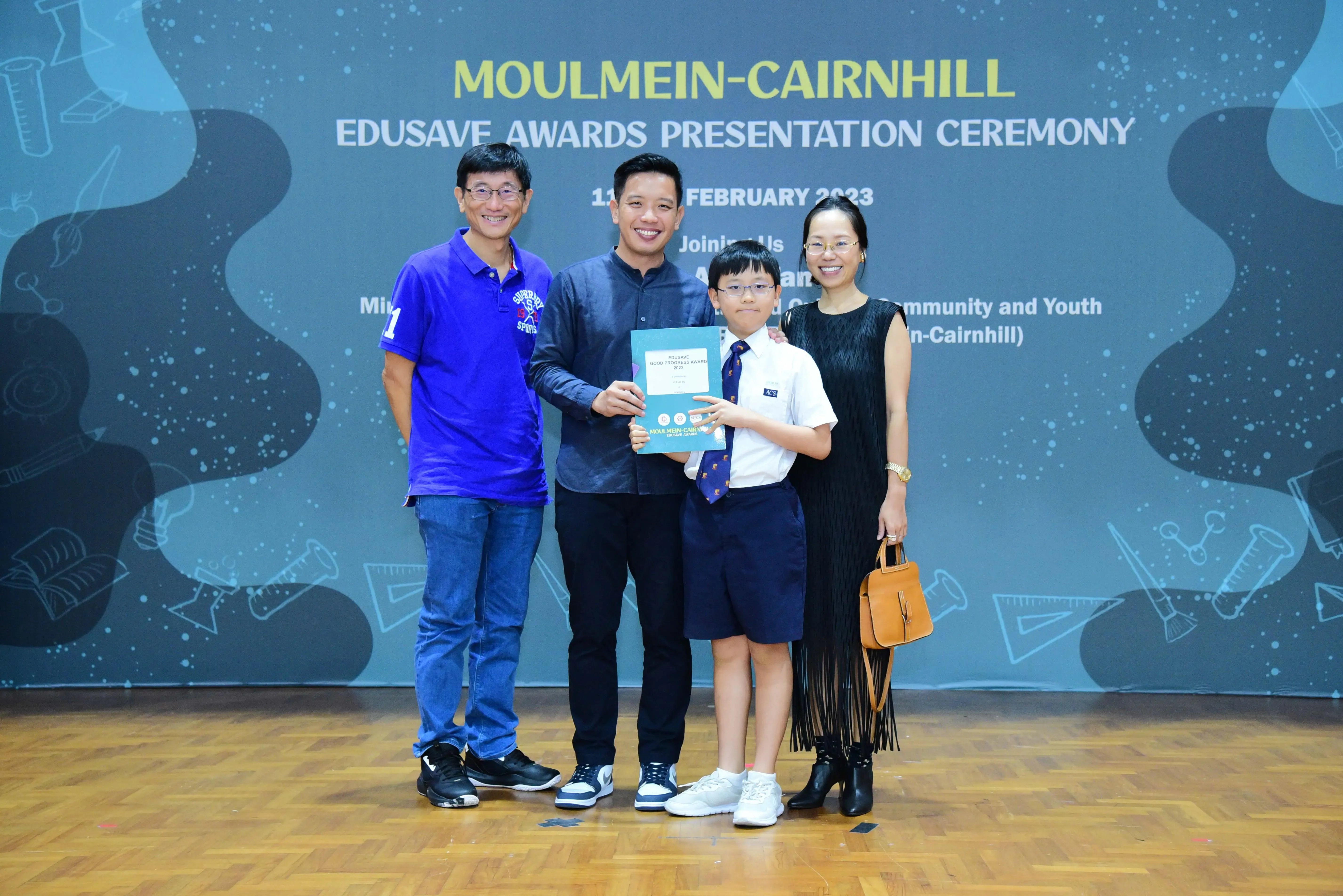 Four people pose at the Moulmein-Cairnhill Edusave Awards Presentation Ceremony, with a boy holding a certificate.