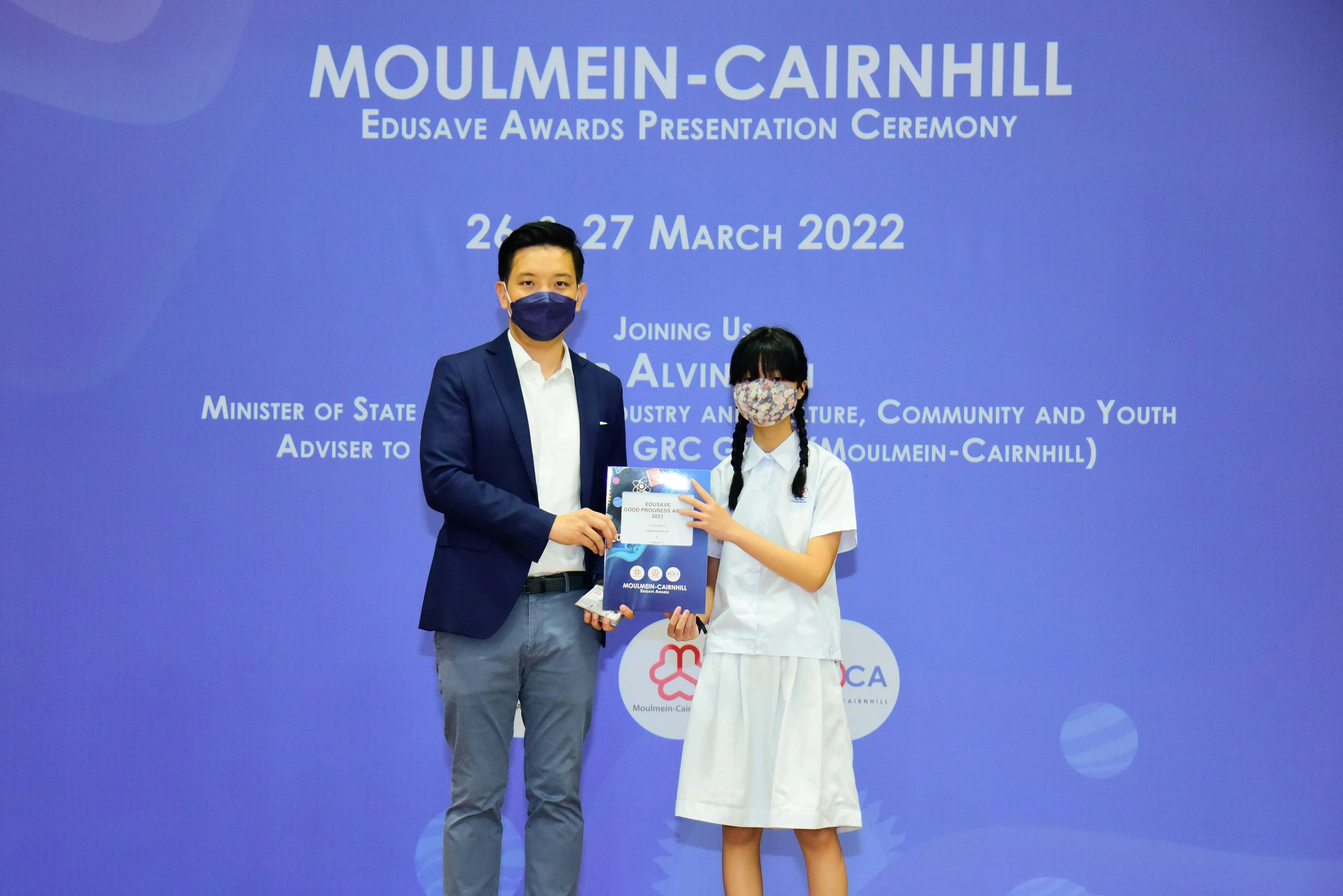 A masked man in a suit and a masked girl in school uniform hold a certificate at the Moulmein-Cairnhill Edusave Awards 2022.