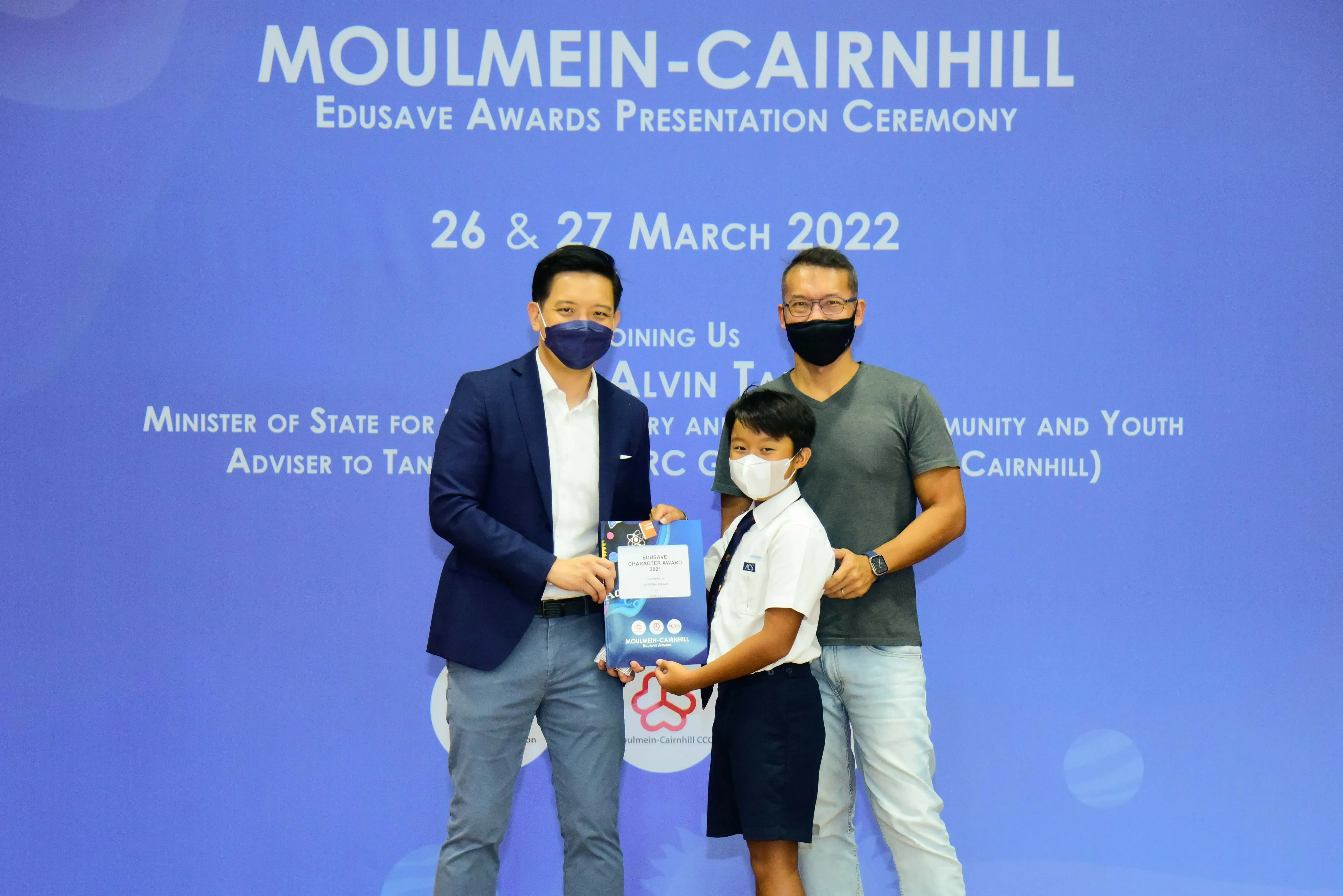 Two masked adults and a uniformed child hold a certificate at the Moulmein-Cairnhill Edusave Awards Ceremony, March 2022.