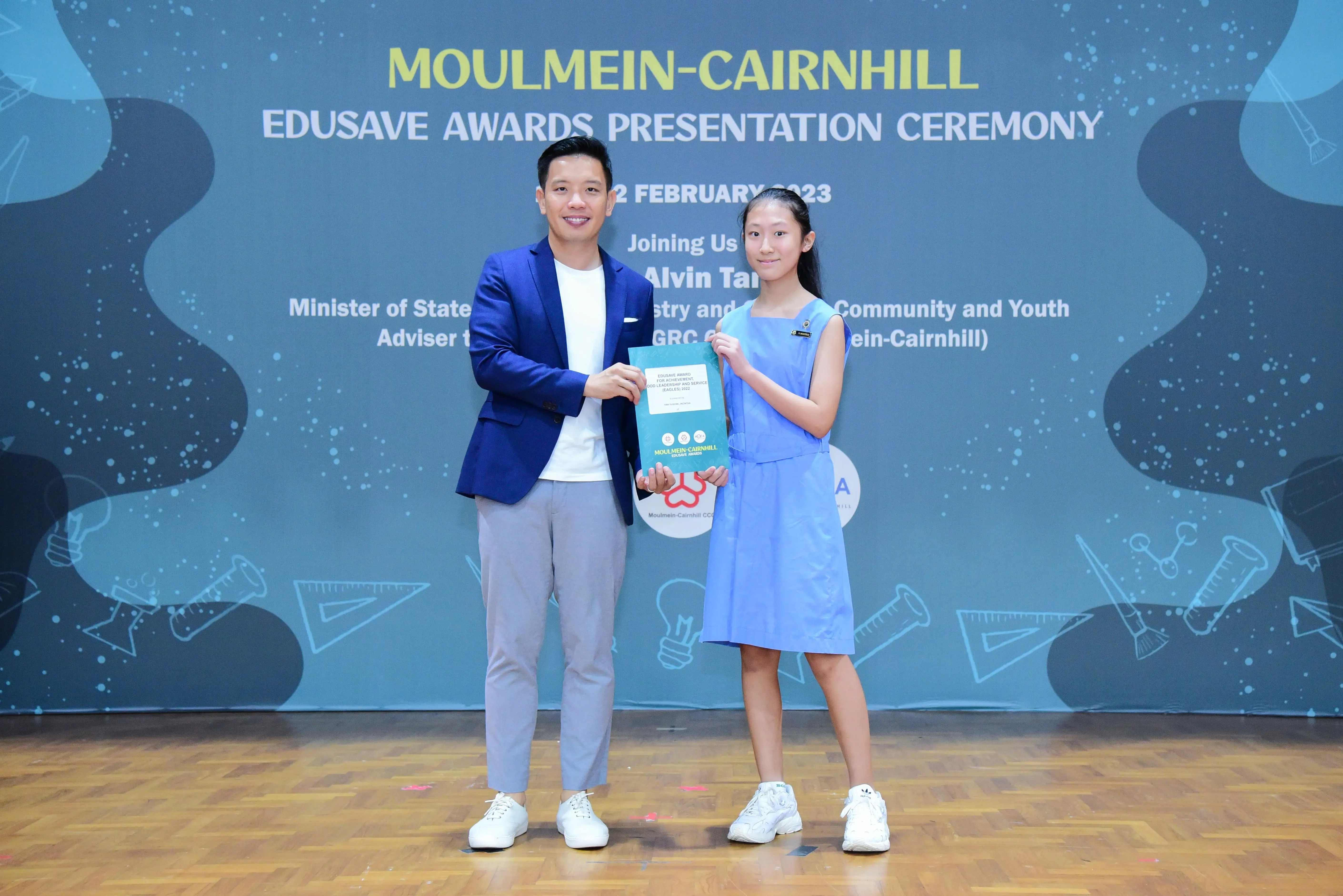 A man in a blue blazer and a young girl in a blue dress hold a certificate at the Moulmein-Cairnhill Edusave Awards Ceremony.