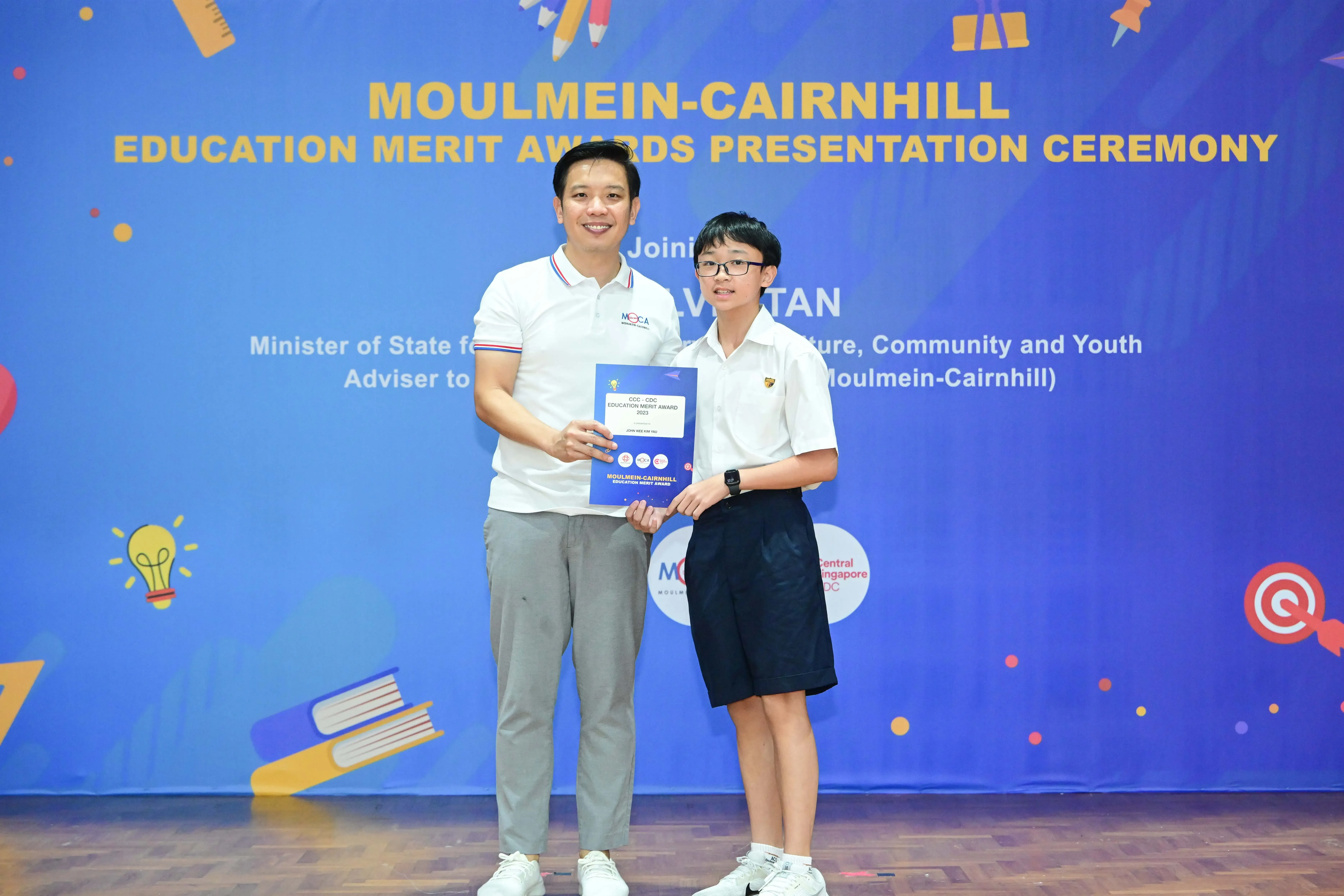 An adult man and a young boy hold an award certificate at the Moulmein-Cairnhill Education Merit Awards ceremony.