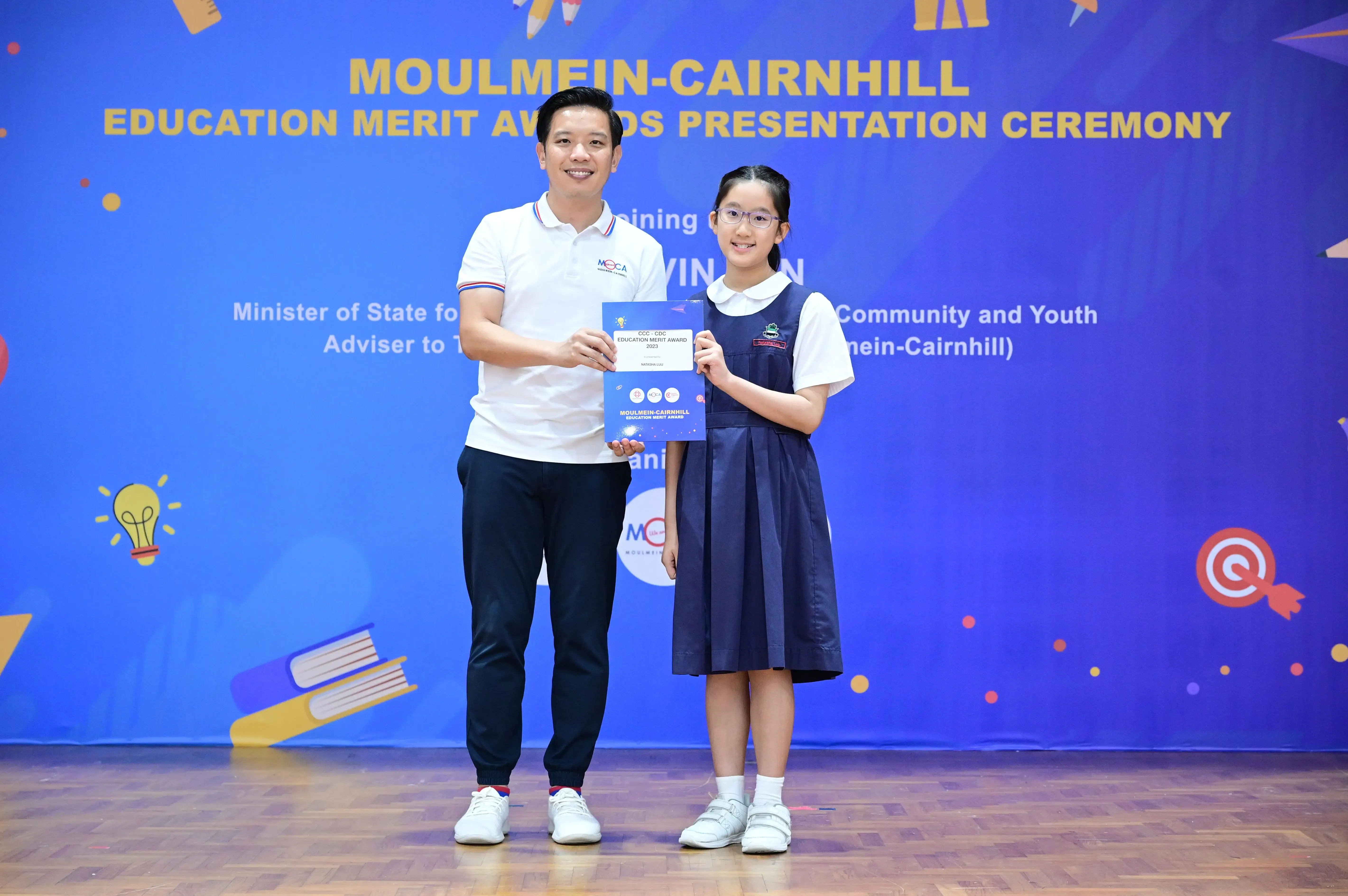 A man and a young girl in school uniform hold an award certificate at the Moulmein-Cairnhill Education Merit Awards ceremony.