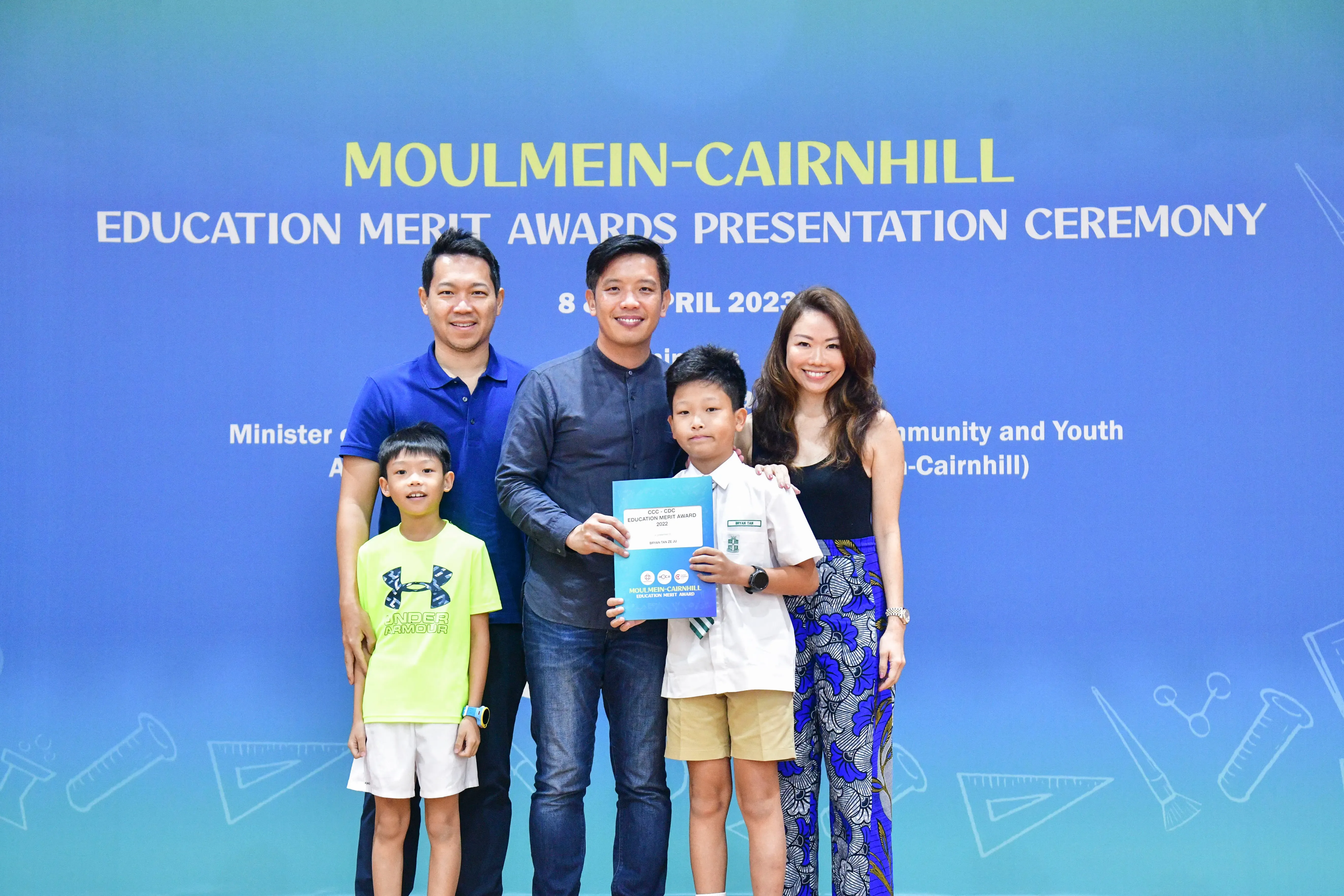 Five people pose before a Moulmein-Cairnhill Education Merit Awards Presentation Ceremony banner on 8 April 2023.