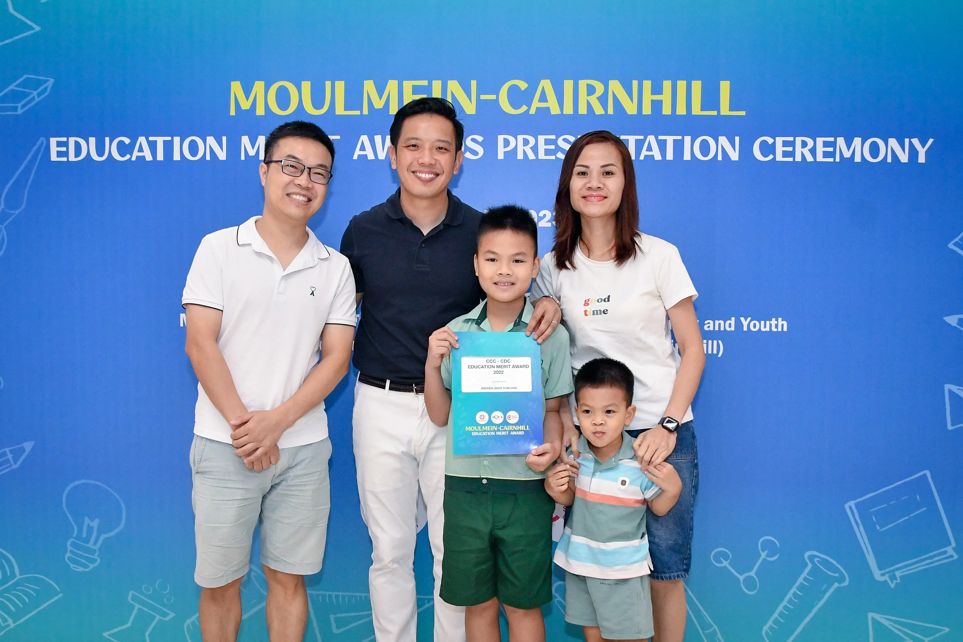 A boy holds a Moulmein-Cairnhill Education Merit Award certificate, posing with three adults and a younger child.