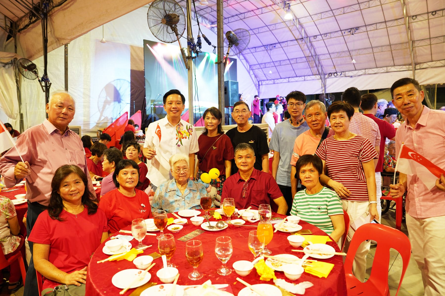 A group of people posing at a red-clothed dinner table under a tent, several holding small Singapore flags.