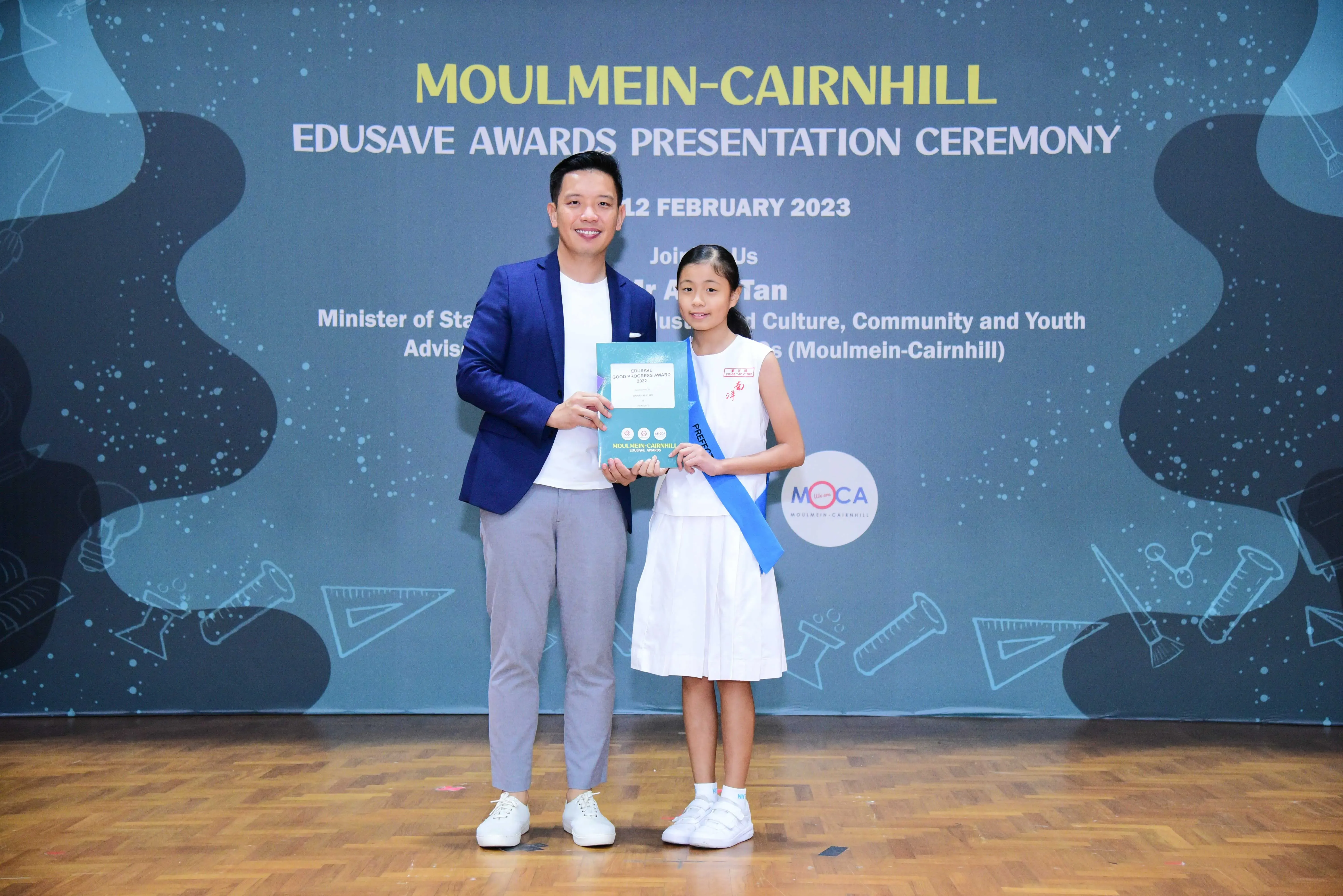 Man in blue blazer and girl in white uniform hold a certificate at Moulmein-Cairnhill Edusave Awards Ceremony 2023.