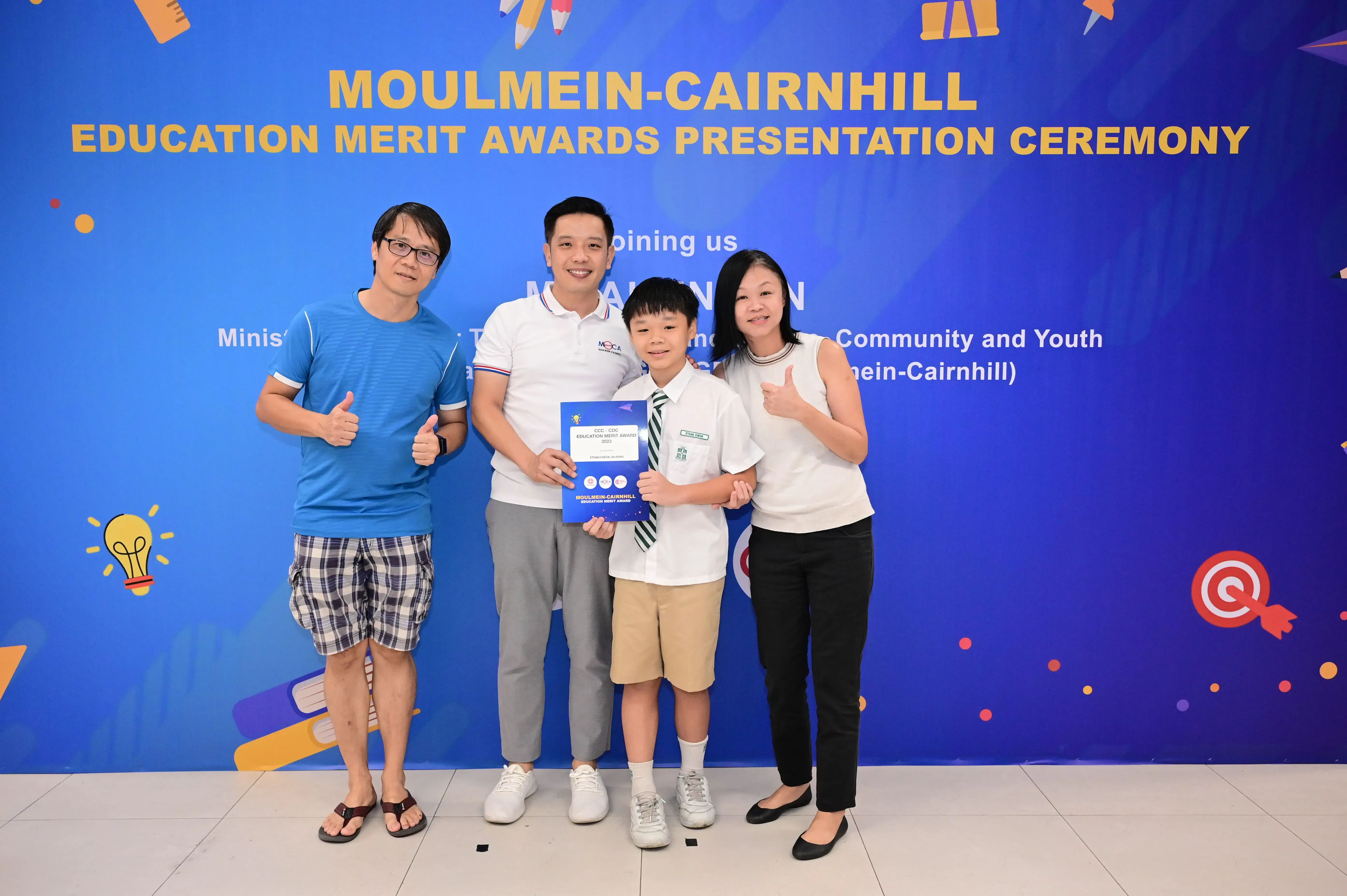 Four people pose before a Moulmein-Cairnhill Education Merit Awards banner, with a boy holding a blue certificate.