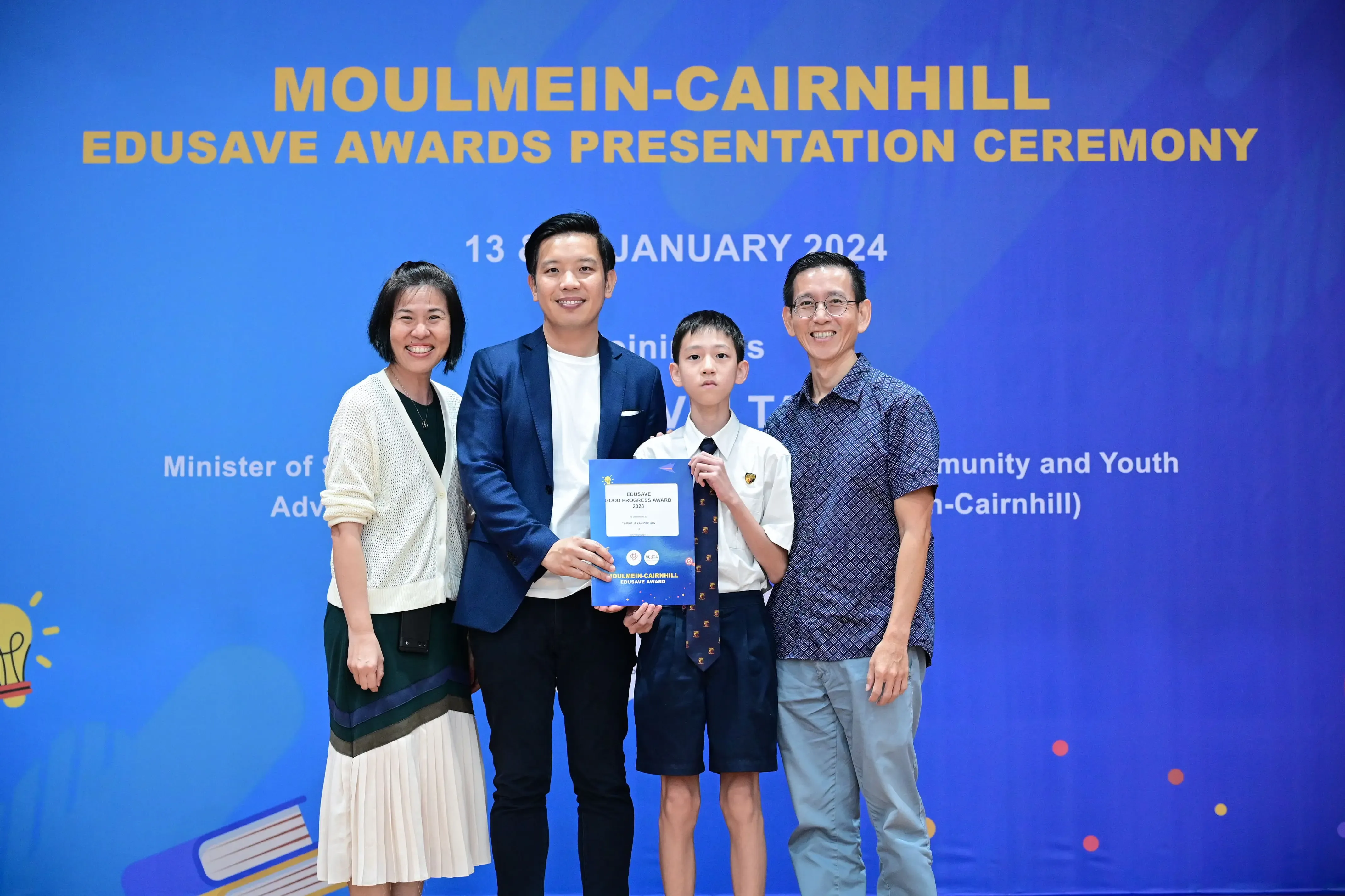 Four people pose at the Moulmein-Cairnhill Edusave Awards Presentation Ceremony, with a boy holding a blue award certificate.