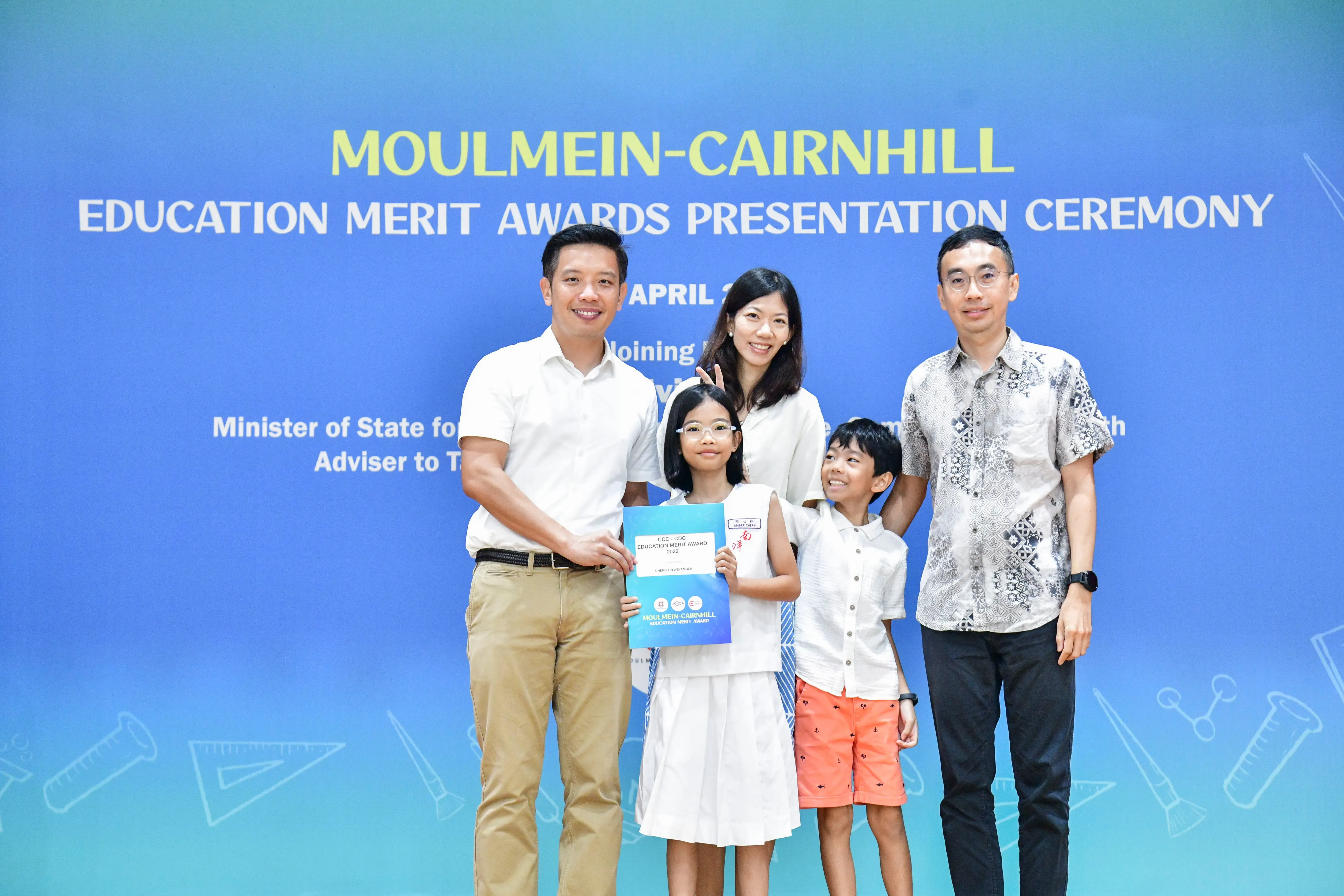 Five people pose before a Moulmein-Cairnhill Education Merit Awards banner, with a girl holding an award certificate.