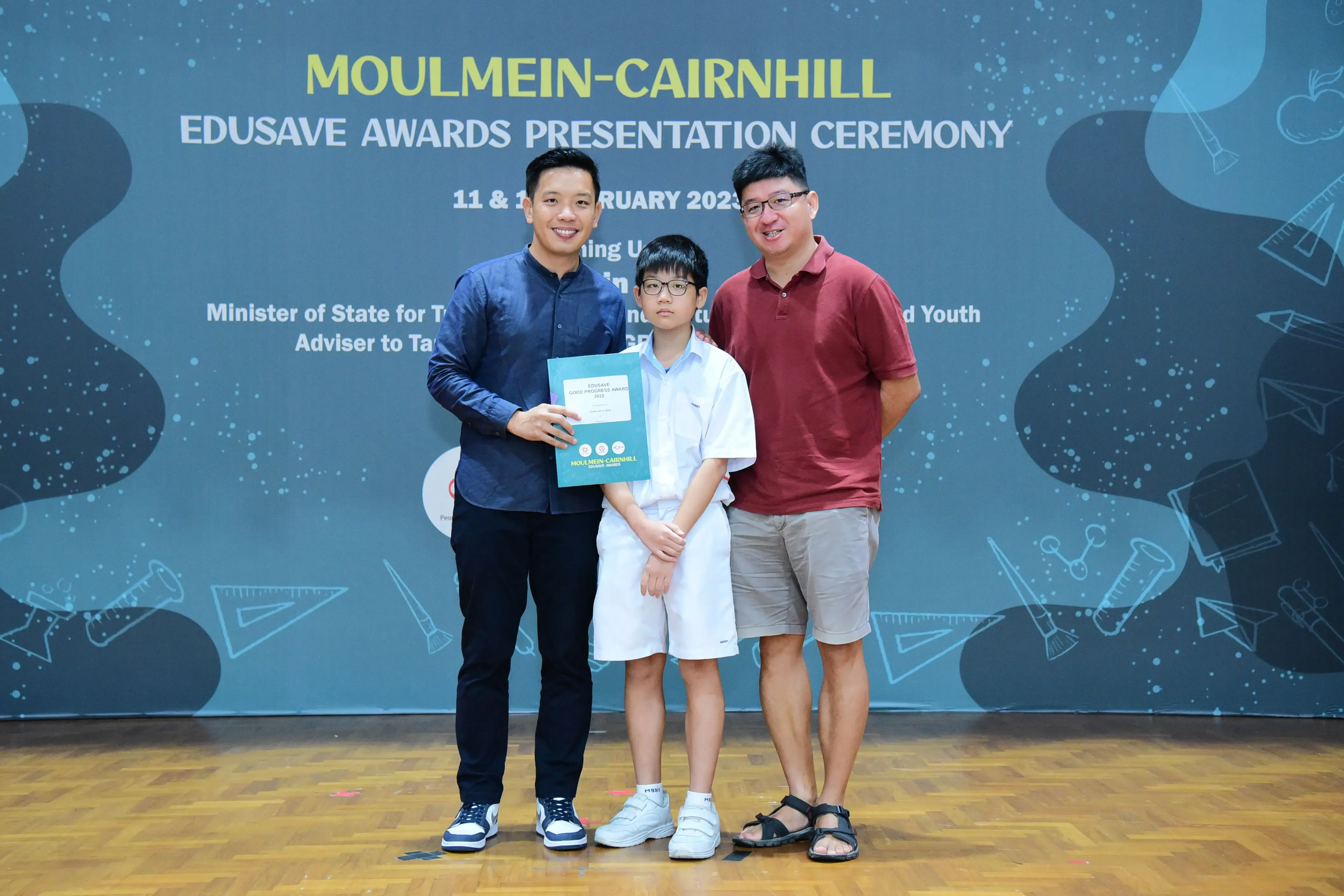 Three people pose at the Moulmein-Cairnhill Edusave Awards Presentation Ceremony 2023, with a boy holding a certificate.