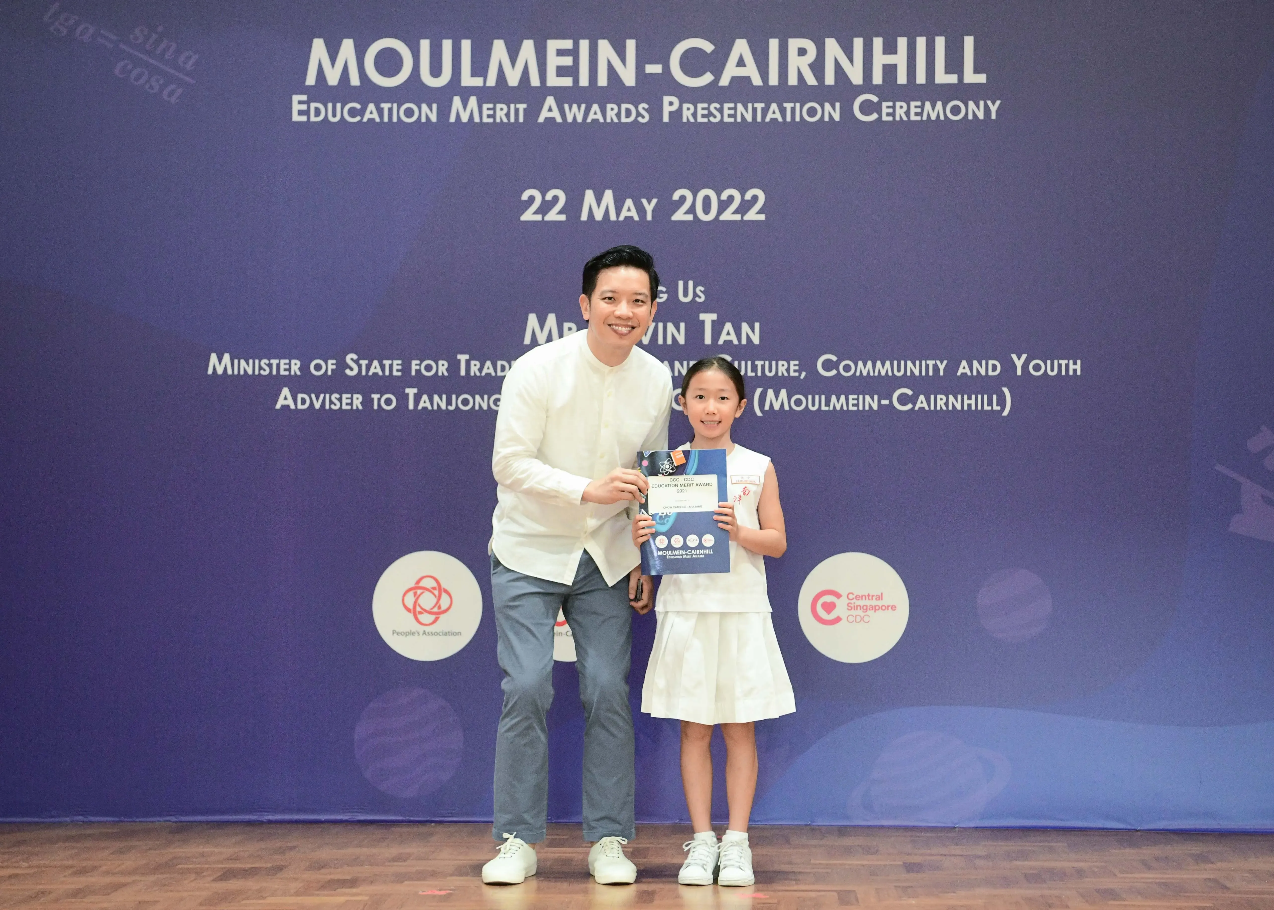 A man and a young girl hold a certificate on stage at the Moulmein-Cairnhill Education Merit Awards on 22 May 2022.