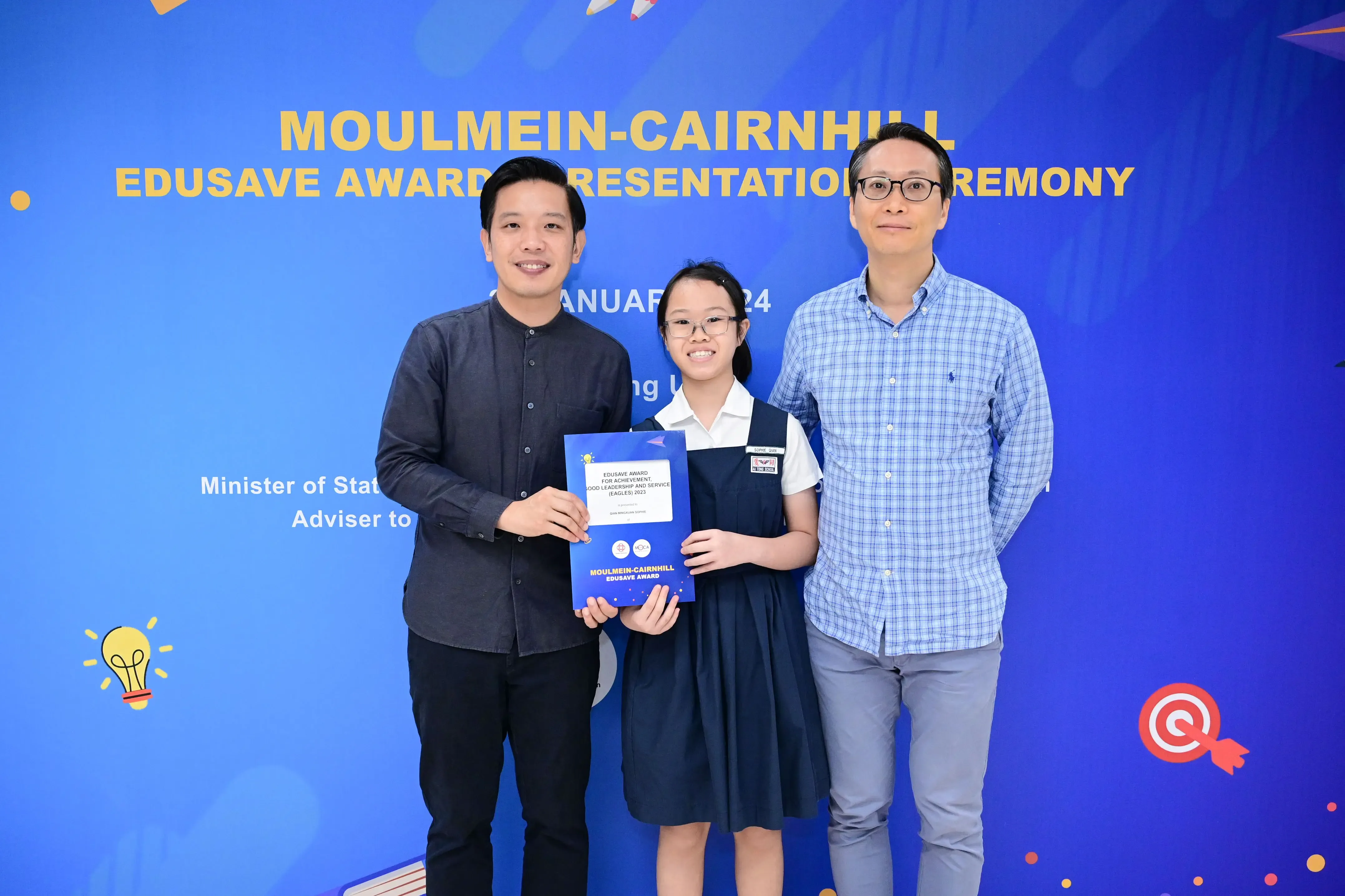 Three people pose before a Moulmein-Cairnhill Edusave Award Presentation Ceremony banner, with a girl holding an award folder.