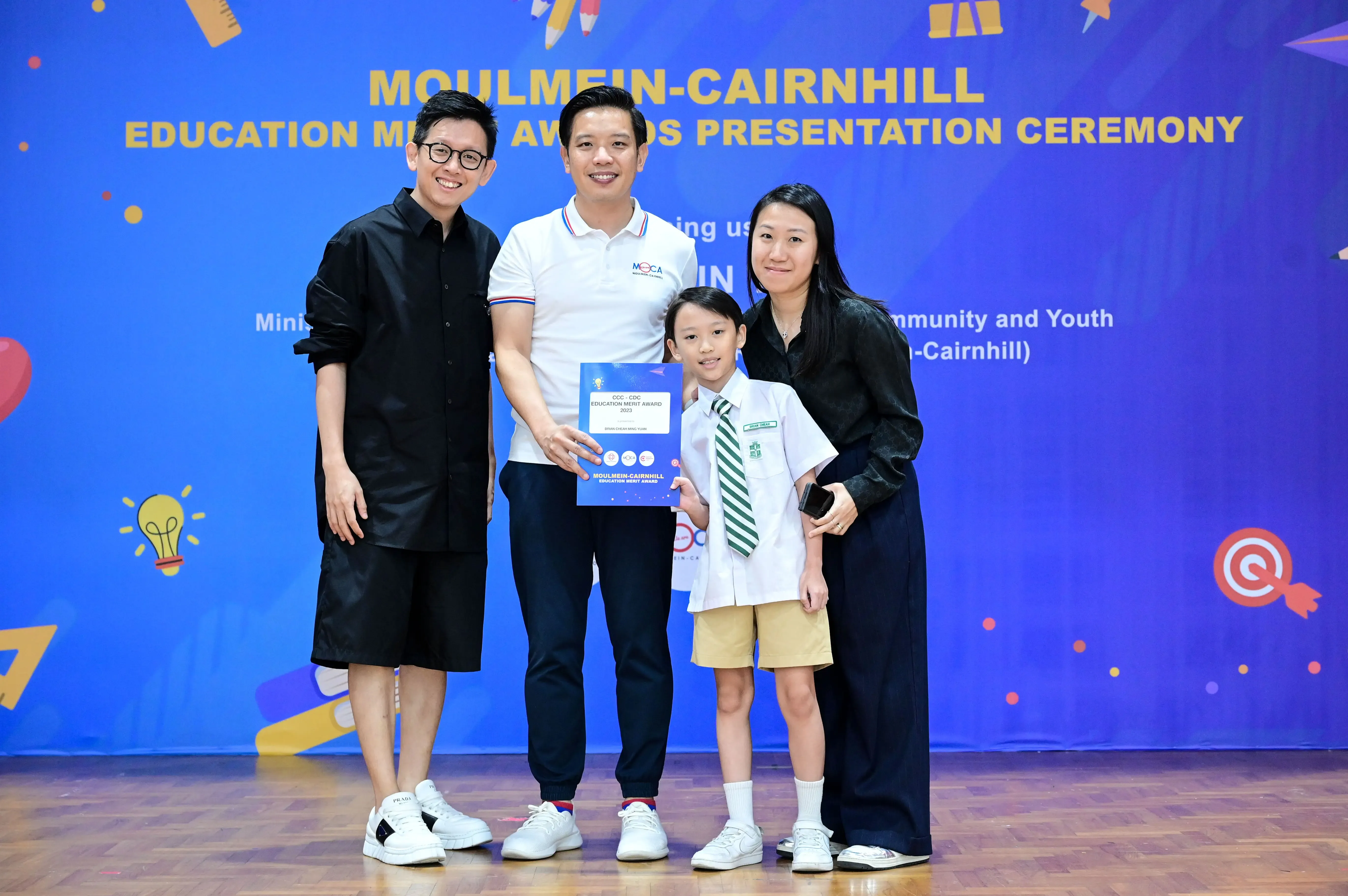 Three adults and a young student in uniform pose with an award certificate at Moulmein-Cairnhill Education Merit Awards ceremony.